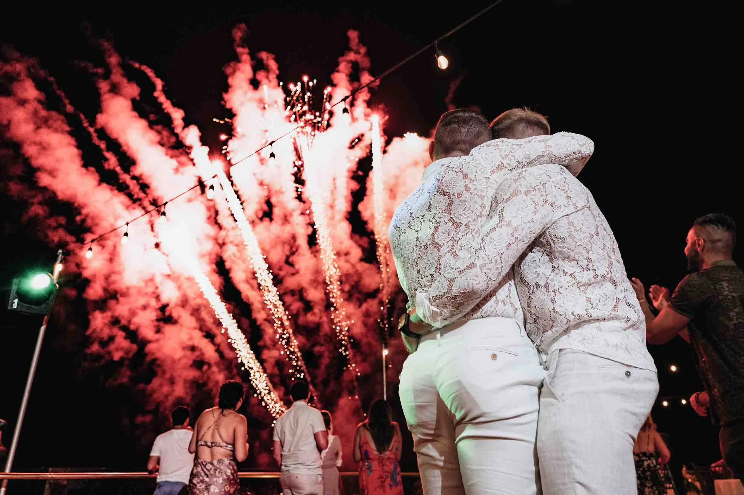 Amazing fireworks during reception of gay wedding, with both grooms in the foreground hugging while they watch them in the sky.
