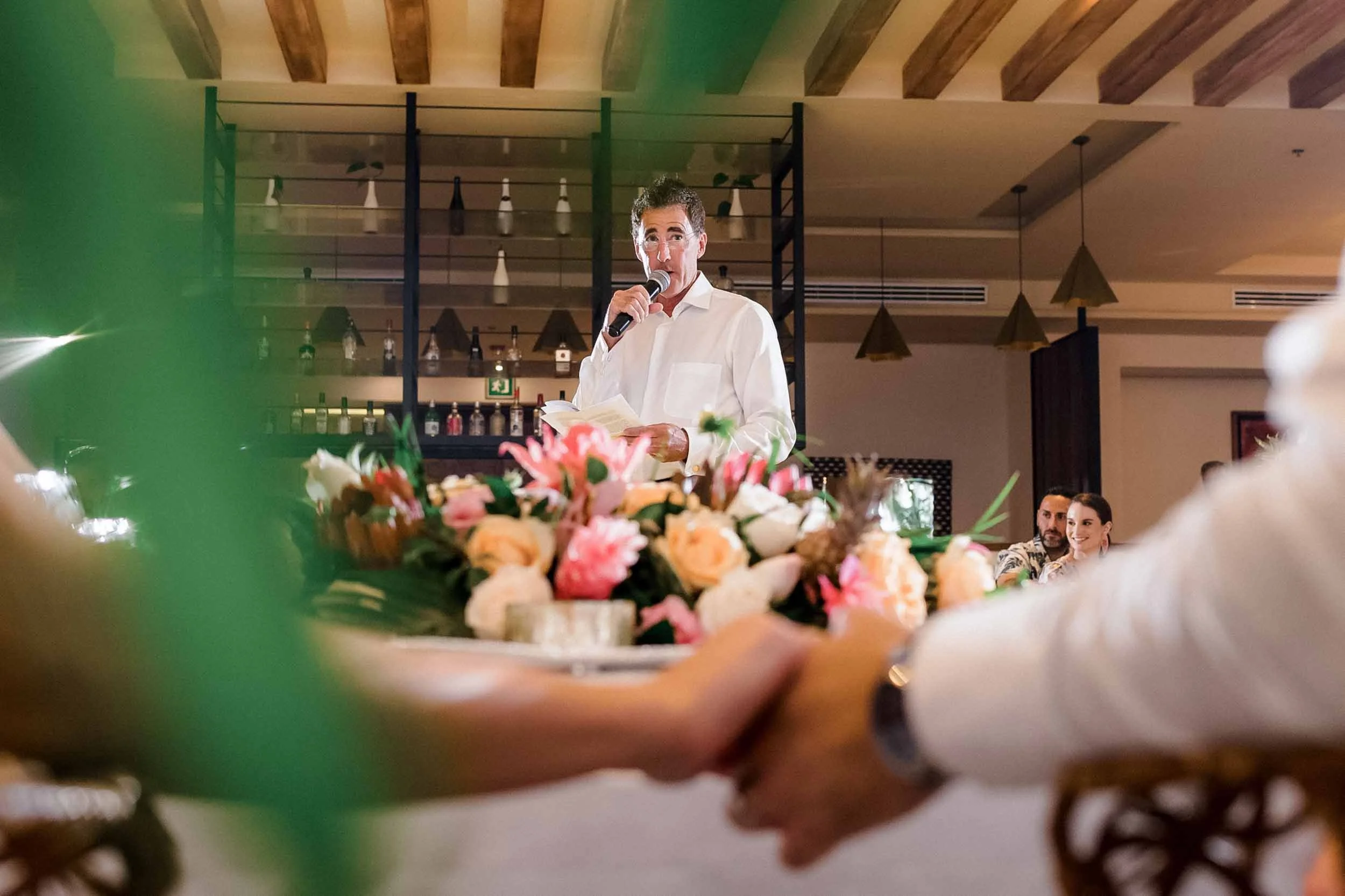 Bride and groom holding hands while man gives speech during wedding reception. 