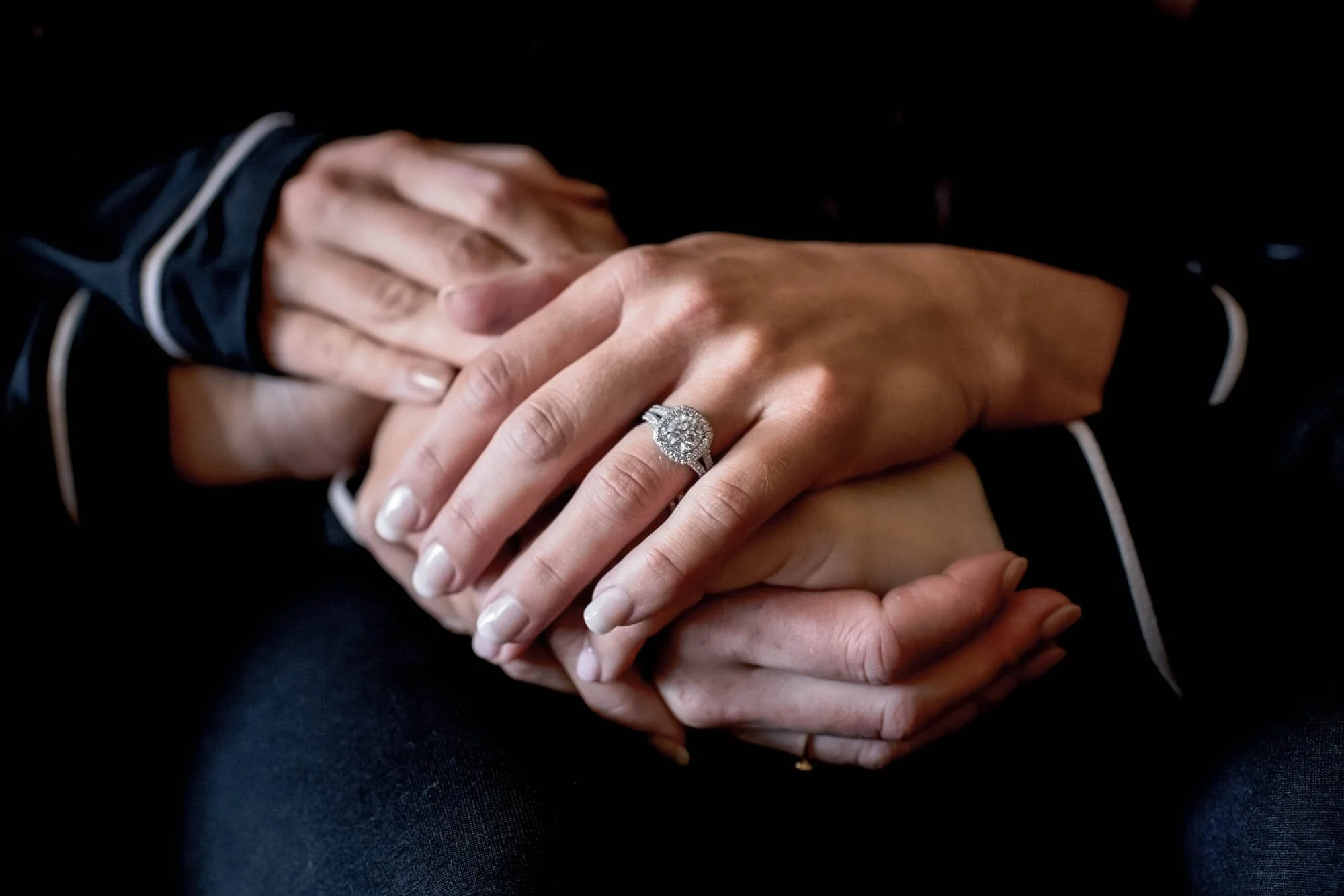 Many hands holding each other, one hand displays a diamond engagement ring
