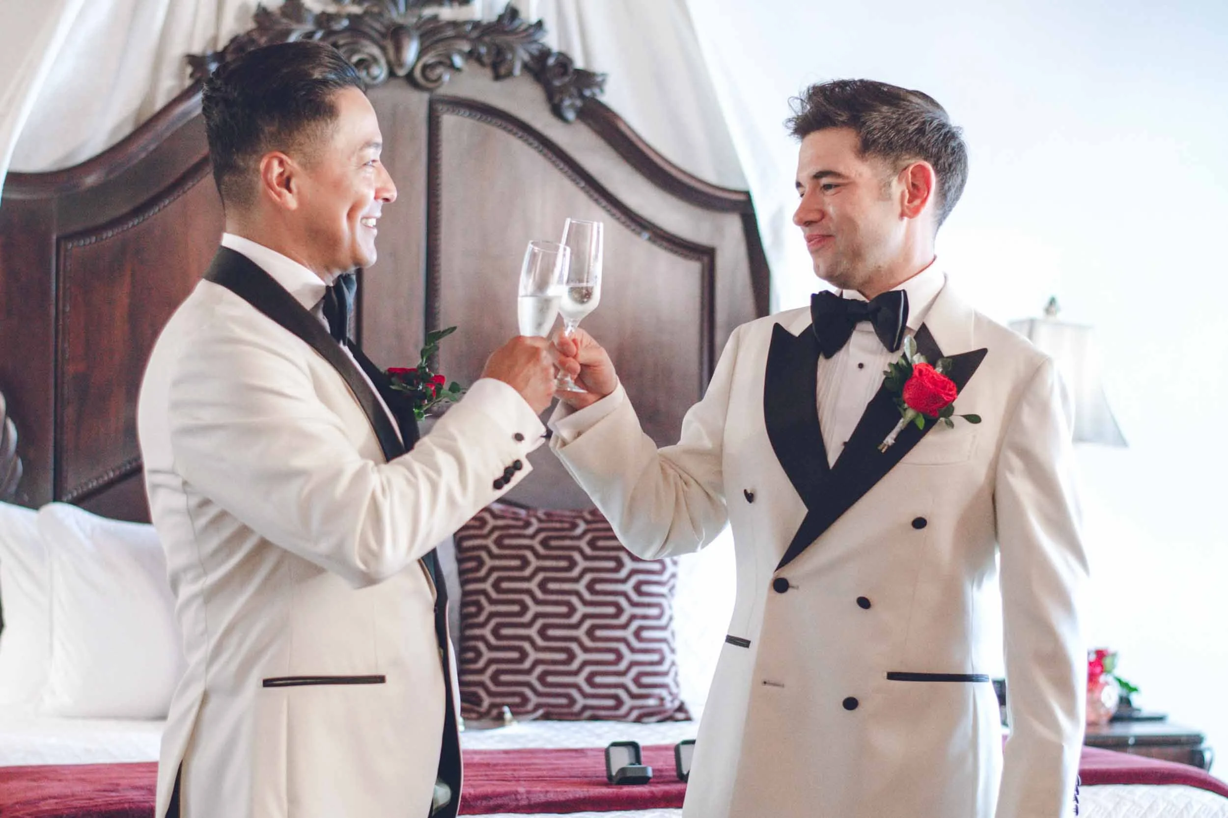 Smiling grooms and clinking champagne glasses, celebrating in a hotel room with ornate headboard and curtains in the background.