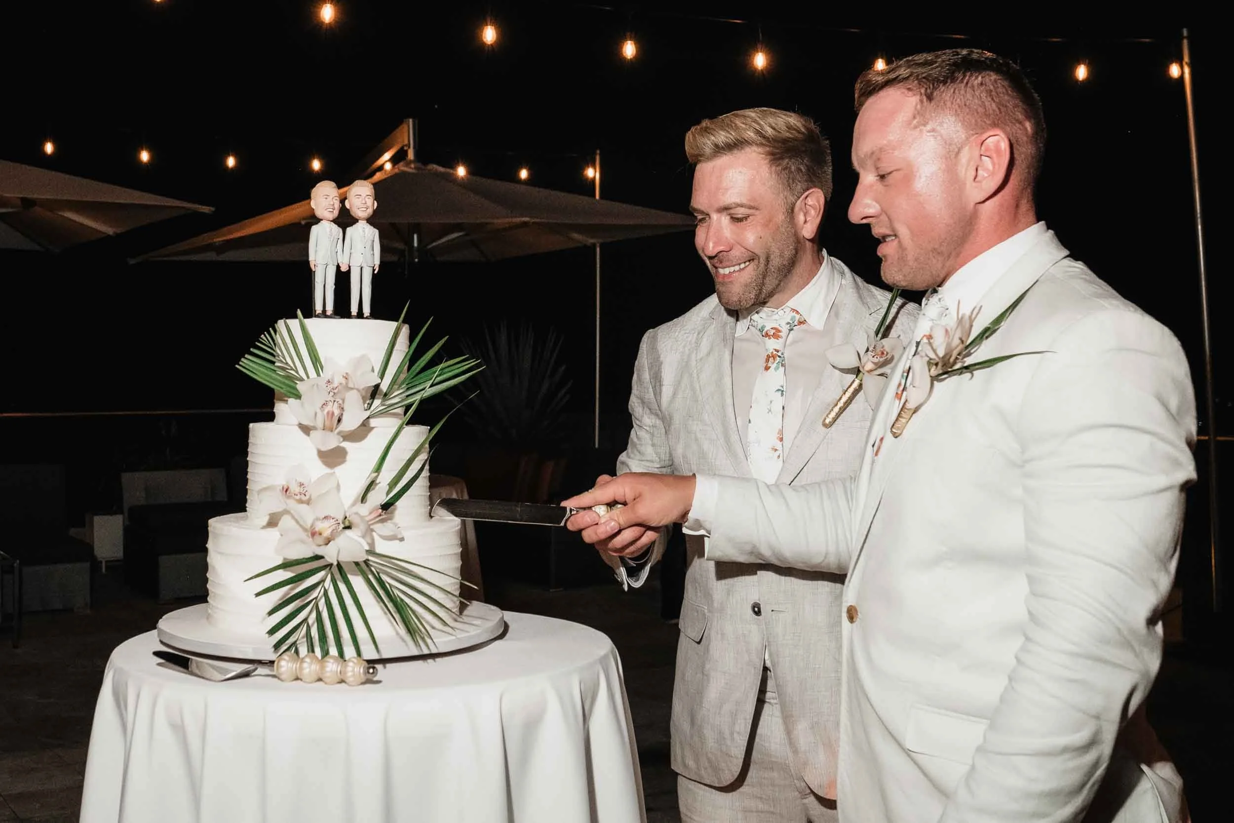 Grooms cutting their  wedding cake with two figurines of them on top, under string lights at night.