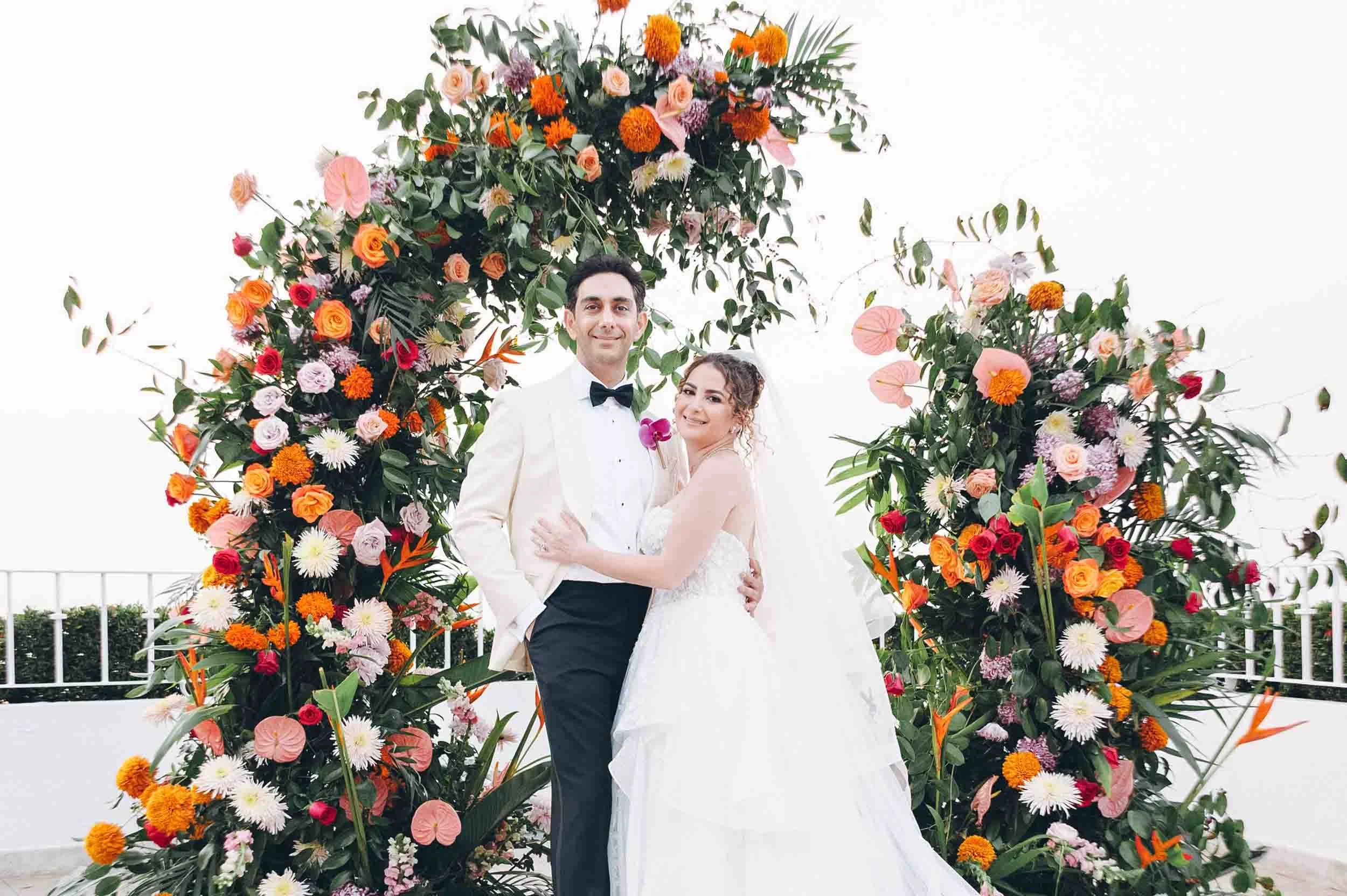 A bride and groom standing together smiling in front of a large floral arch with pink, orange, and white flowers.