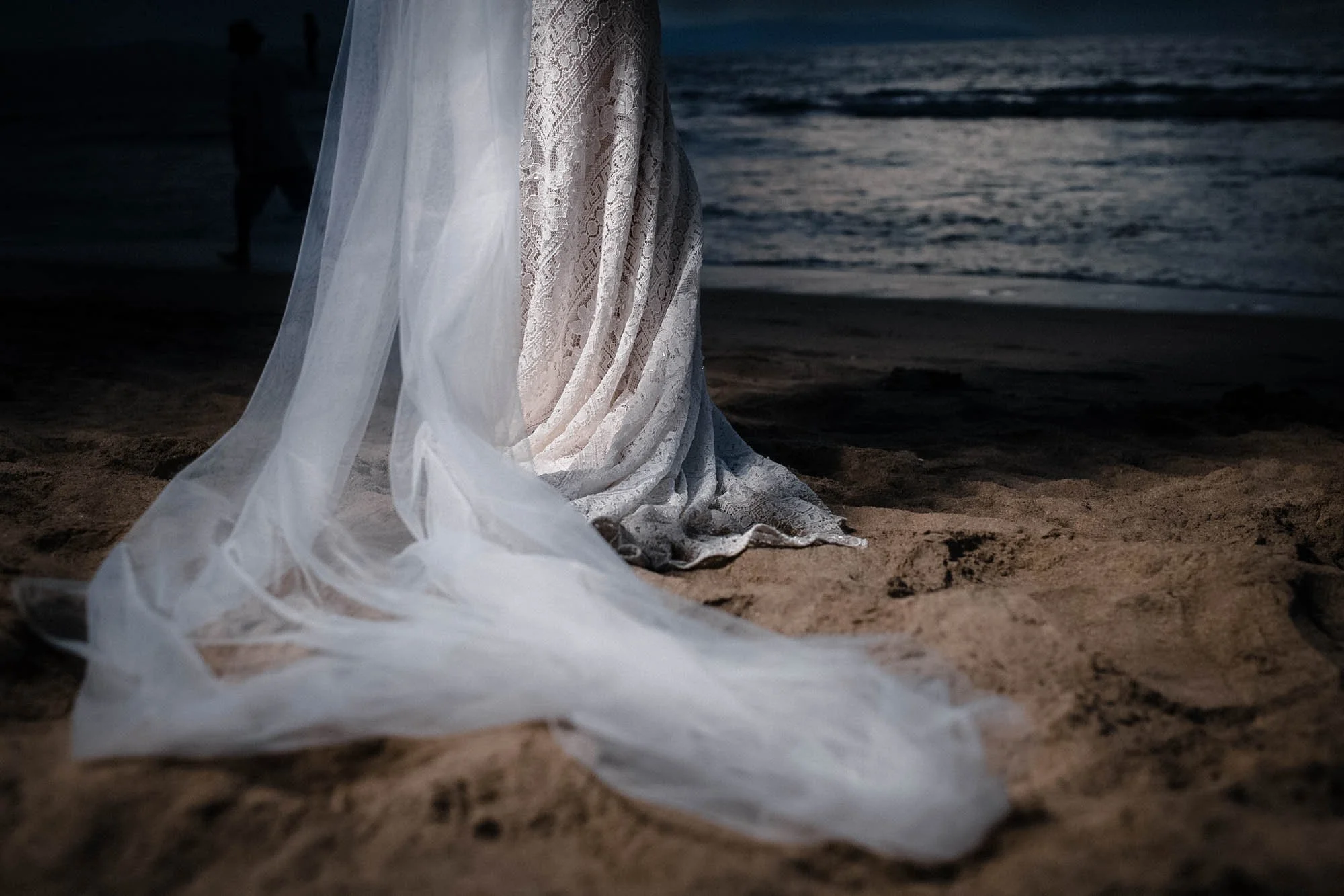Veil and dress detail on sand and ocean shore in the background.