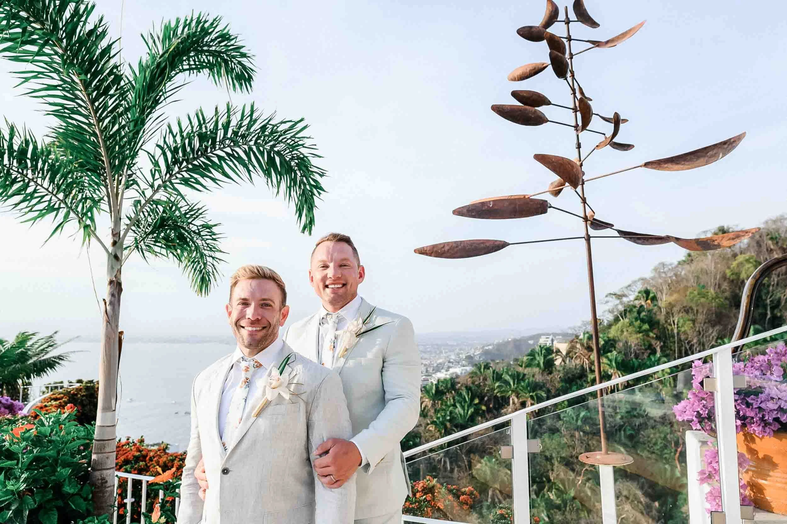 Gay couple posing after their outdoor wedding ceremony with greenery and flowers around, overlooking the ocean from a hillside in the background from the Grand Miramar Luxury resort.