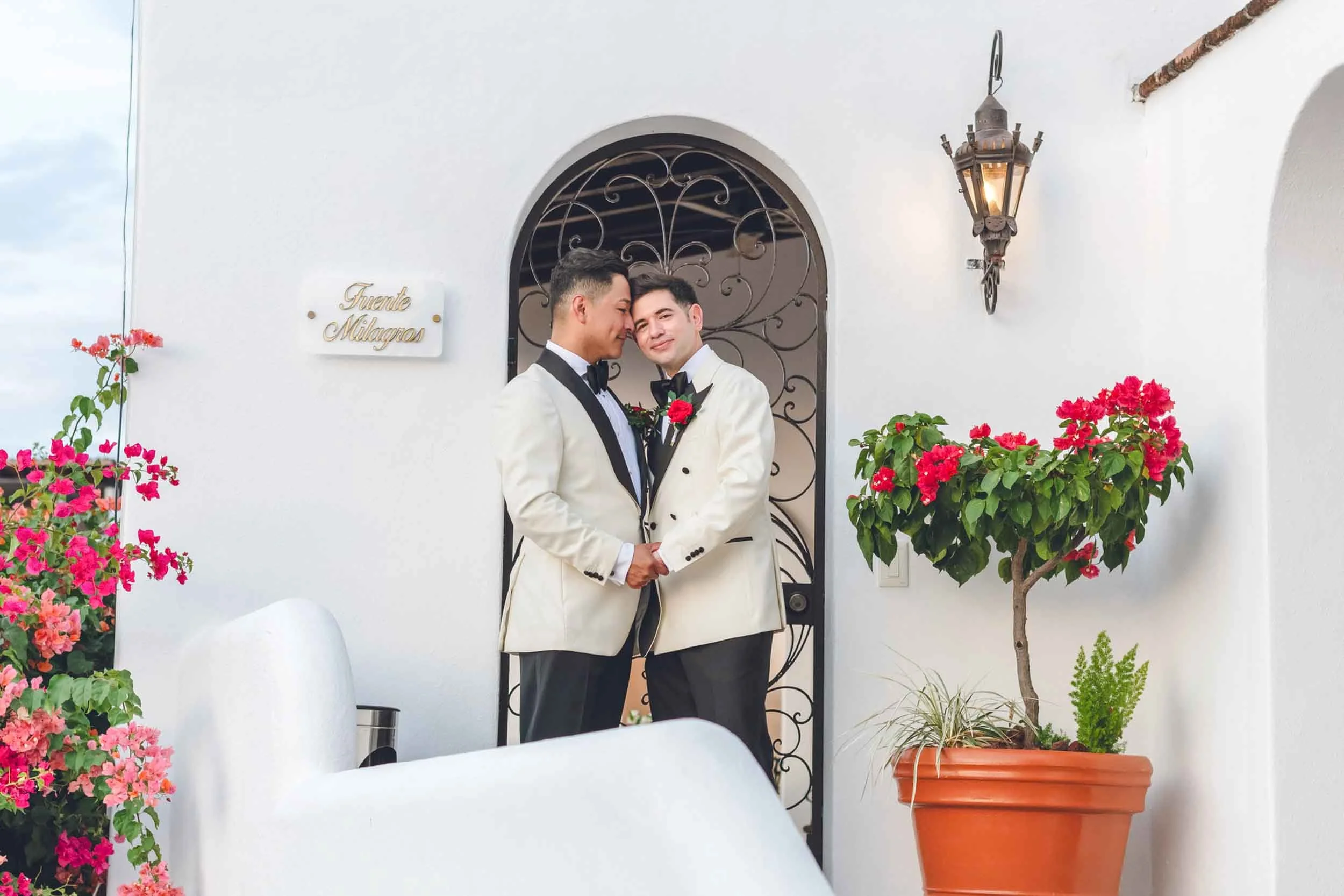 Wedding portrait of gay couple tuxedos holding hands and leaning their foreheads together in front of a white wall with flowers and a lantern.