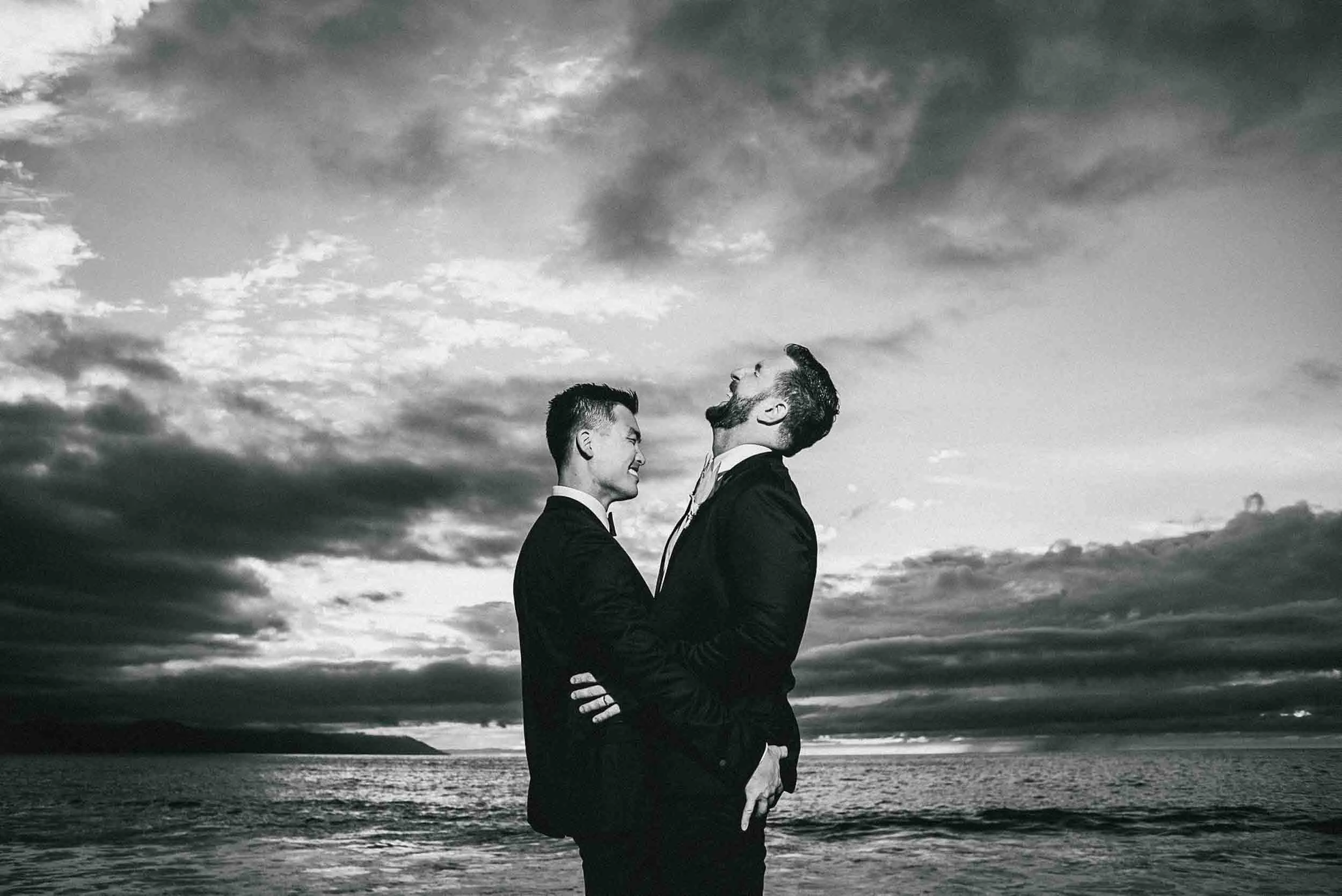 Grooms hugging face to face on a beach with the ocean and cloudy sky in the background, smiling and looking at each other.