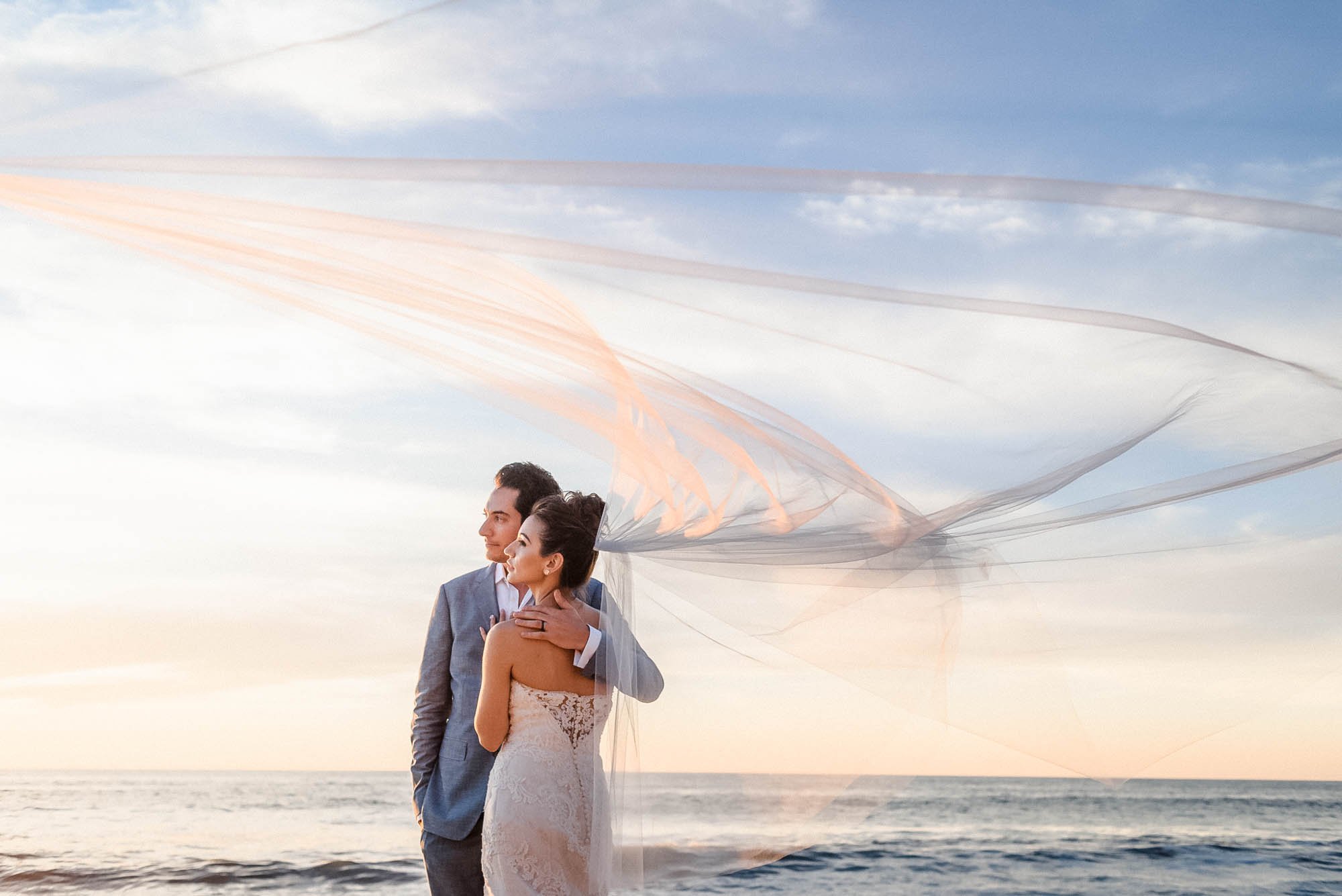 Bride and groom posing on beach with flying veil and pastel sunset.
