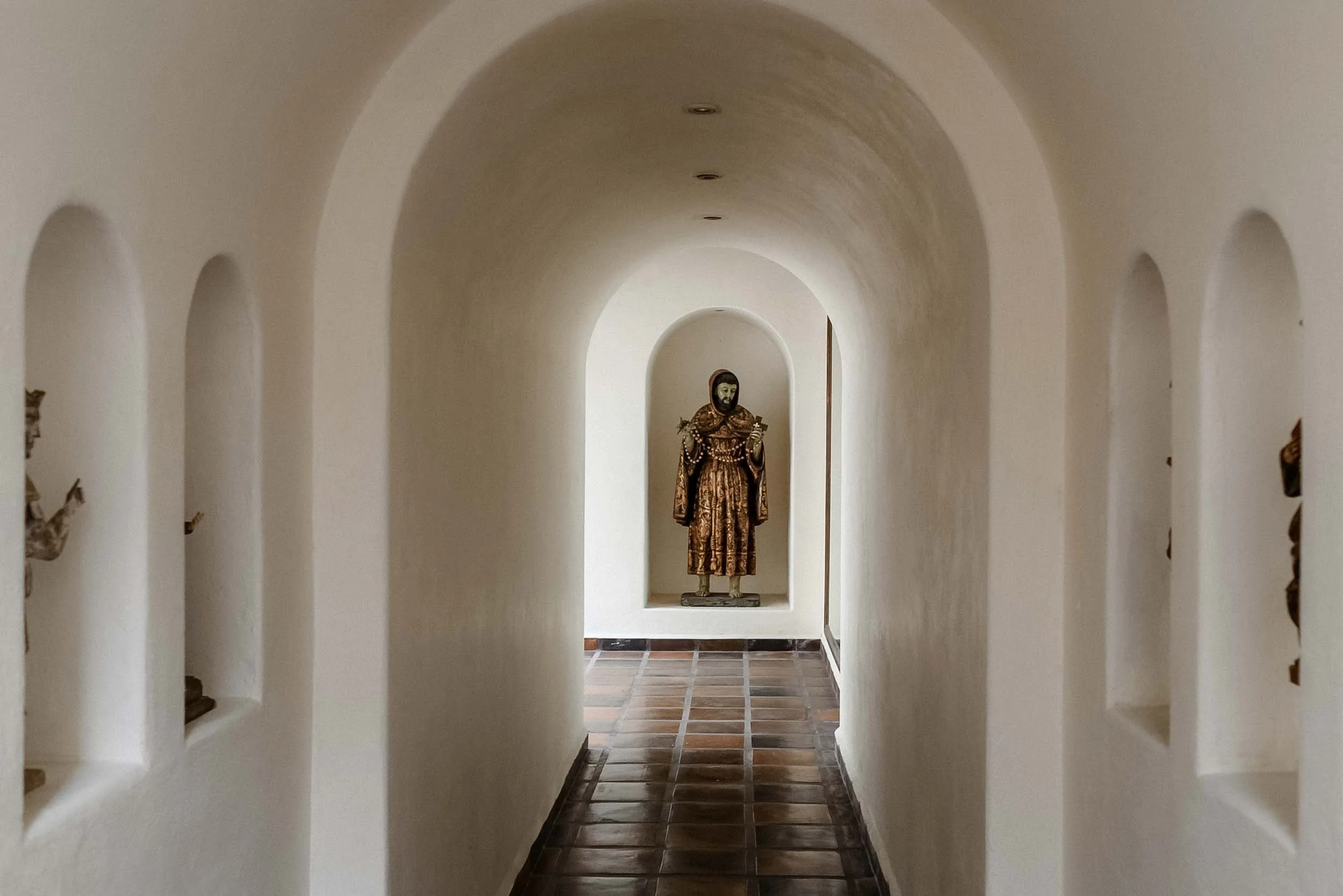 A hallway with rounded arches on both sides leading to a statue of a religious figure at the end.