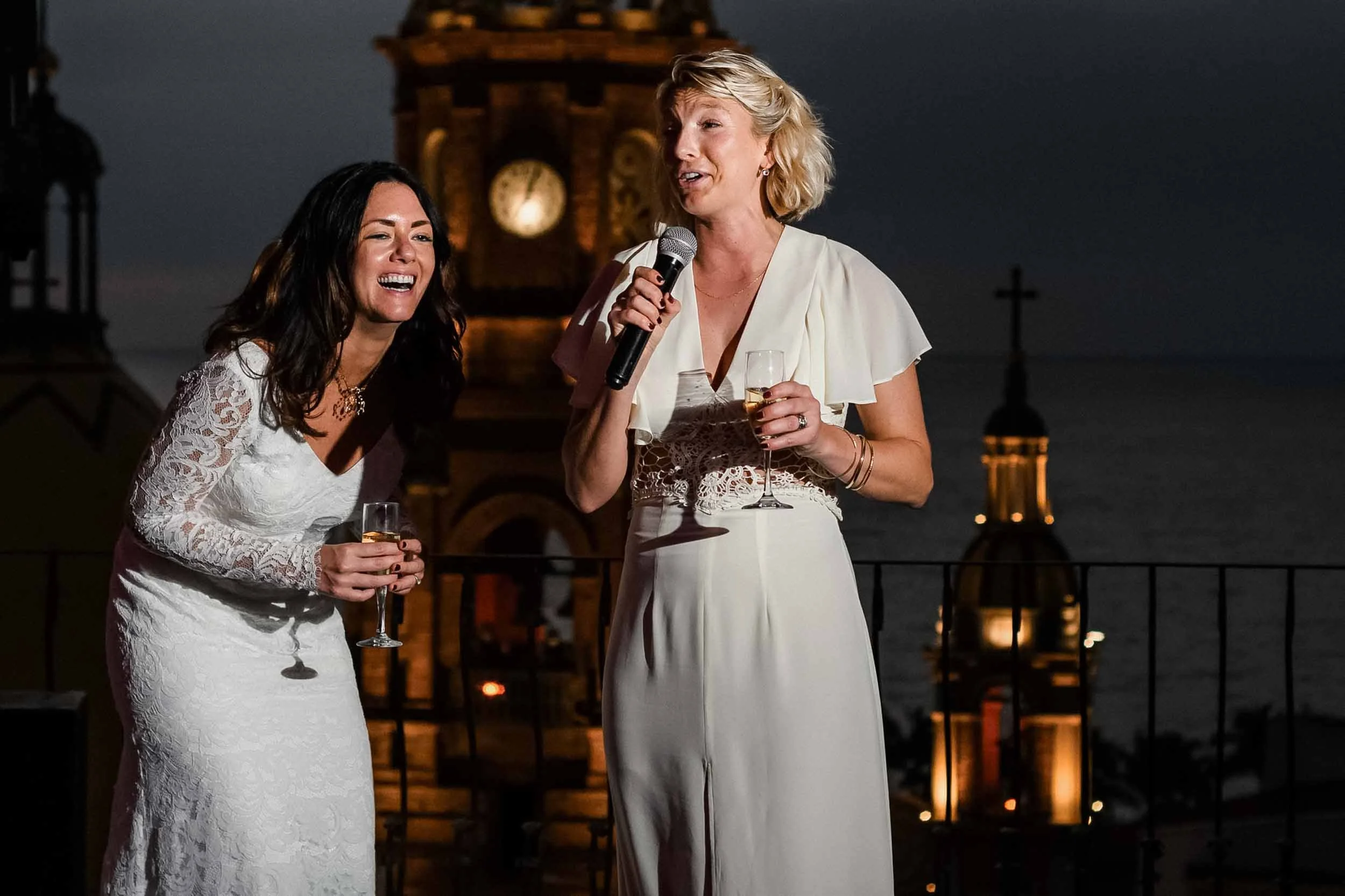 Bride and maid of honor holding glasses of champagne while giving speeches at wedding reception, with a historic clock tower and dark sky in the background.