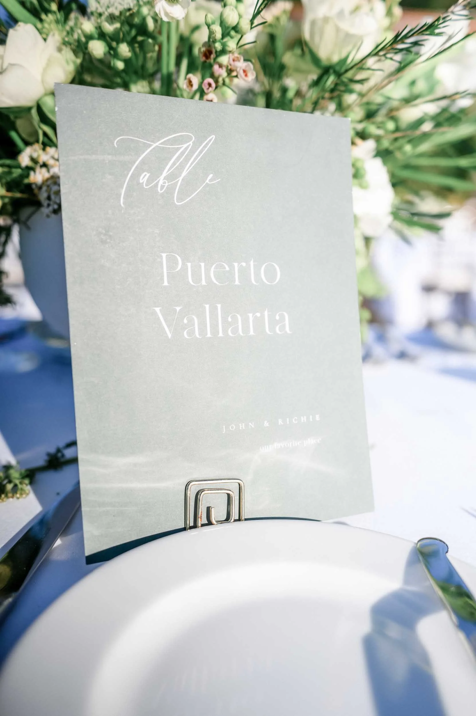 A card with the words 'Table Puerto Vallarta' printed on it, attached to a stand inside a setting with a floral arrangement in the background.