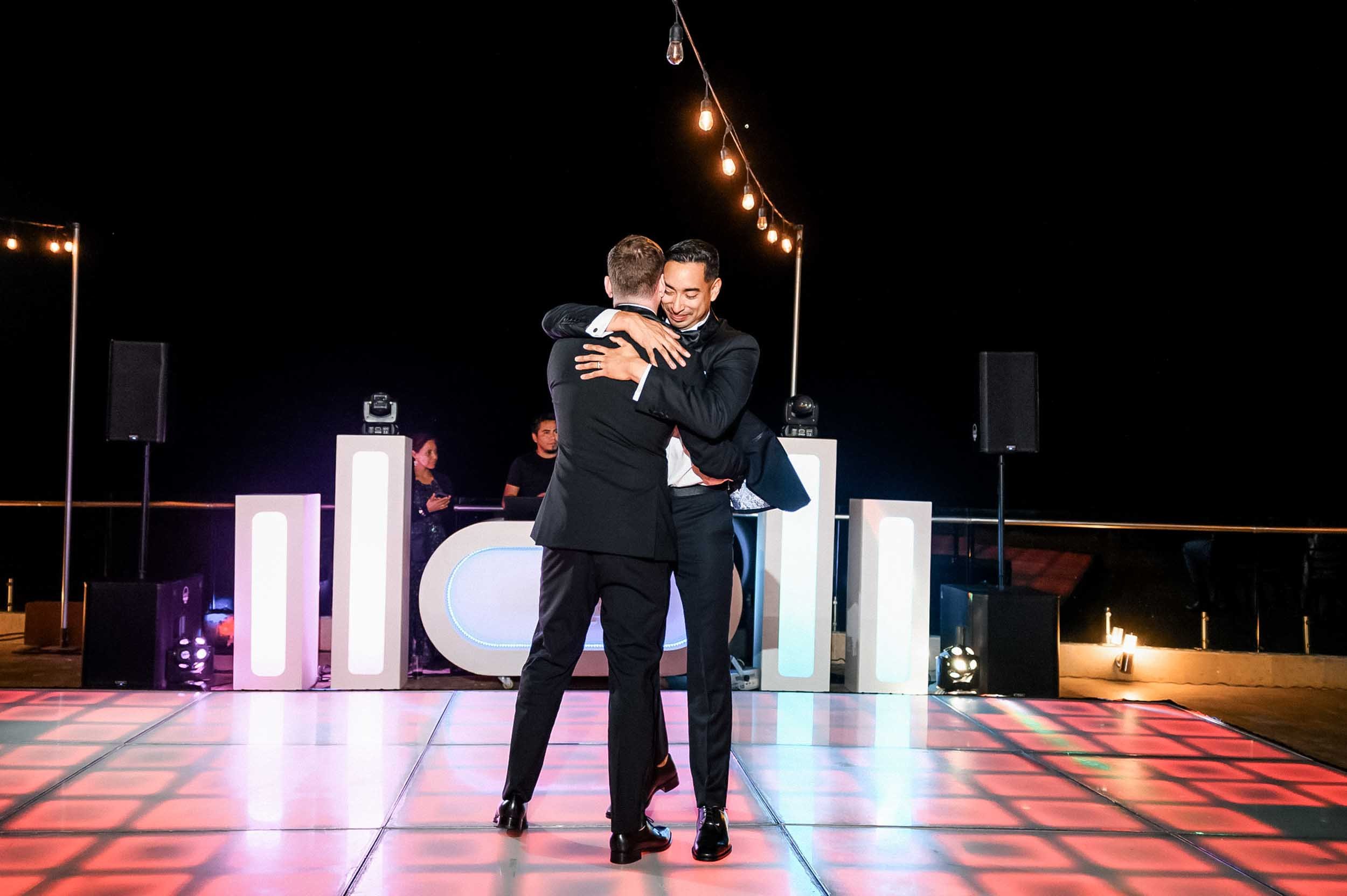 Grooms dancing closely during their first dance, with a black background and string lights overhead, and a DJ booth in the background.