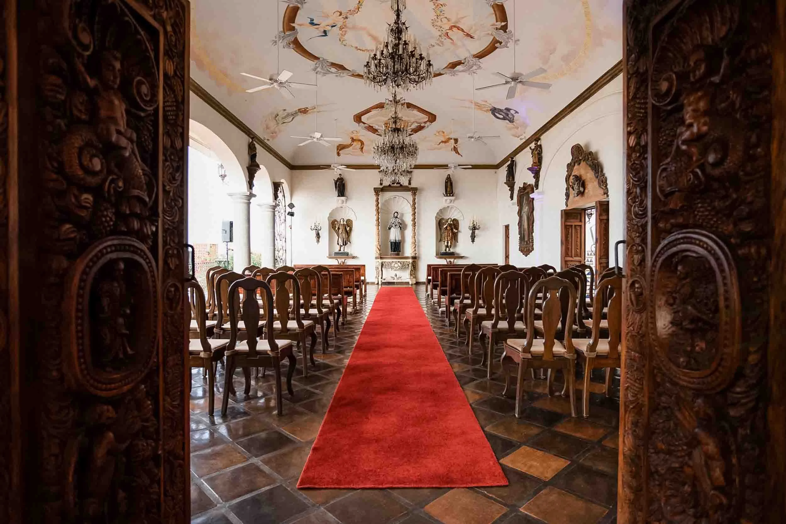 View of La Capella wedding venue with carved wooden doors, a red carpet aisle, statues, chandeliers, and a painted ceiling with angels.