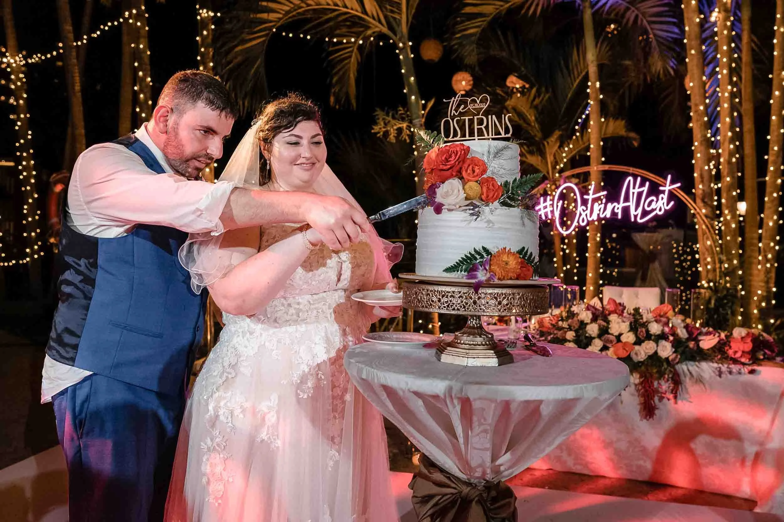 Bride and groom cutting wedding cake at outdoor reception with string lights and palm trees in background.