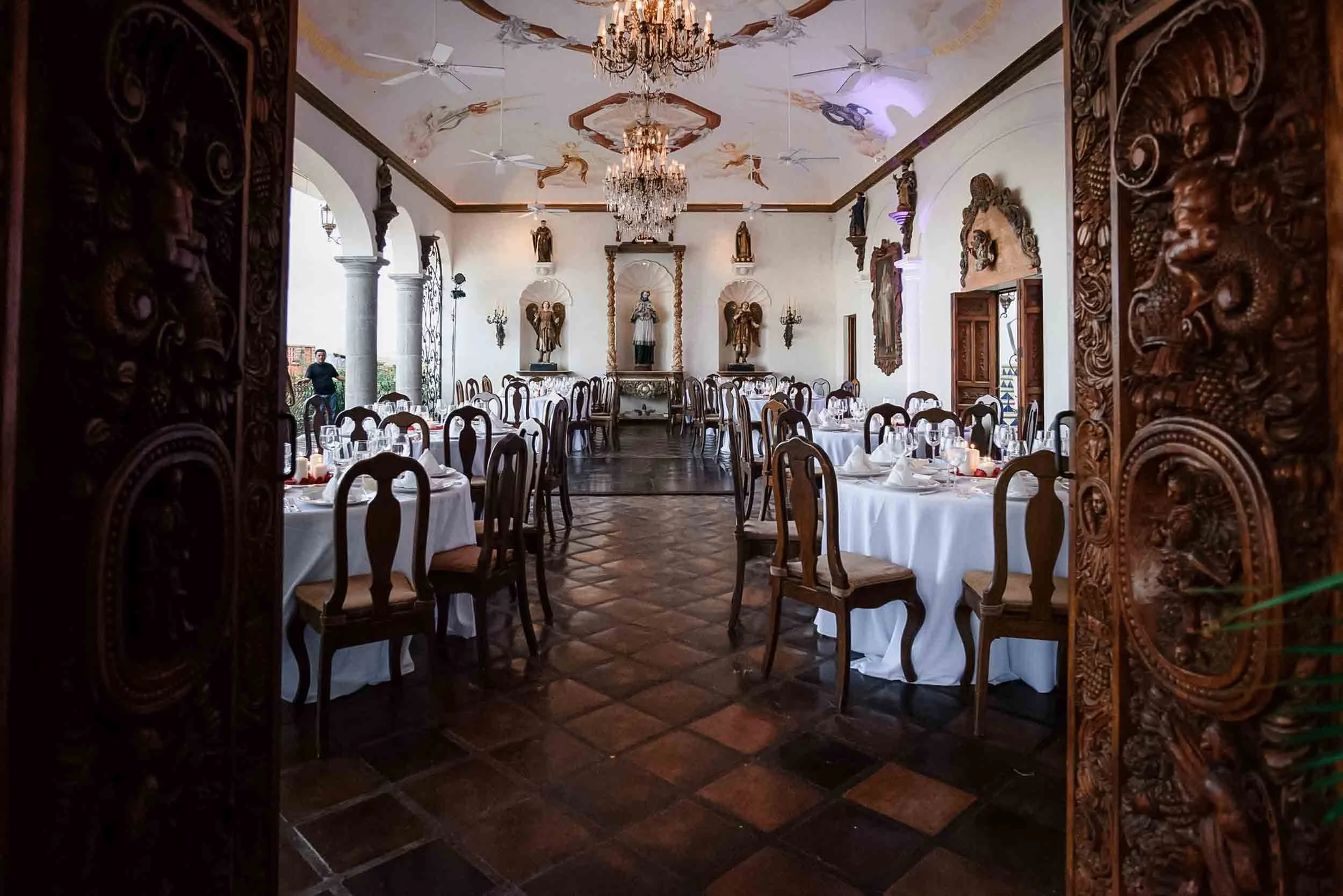  Luxurious La Capella interior decorated with religious statues, chandeliers, and ornate wood carvings, set with round tables covered in white tablecloths and elegant table settings.