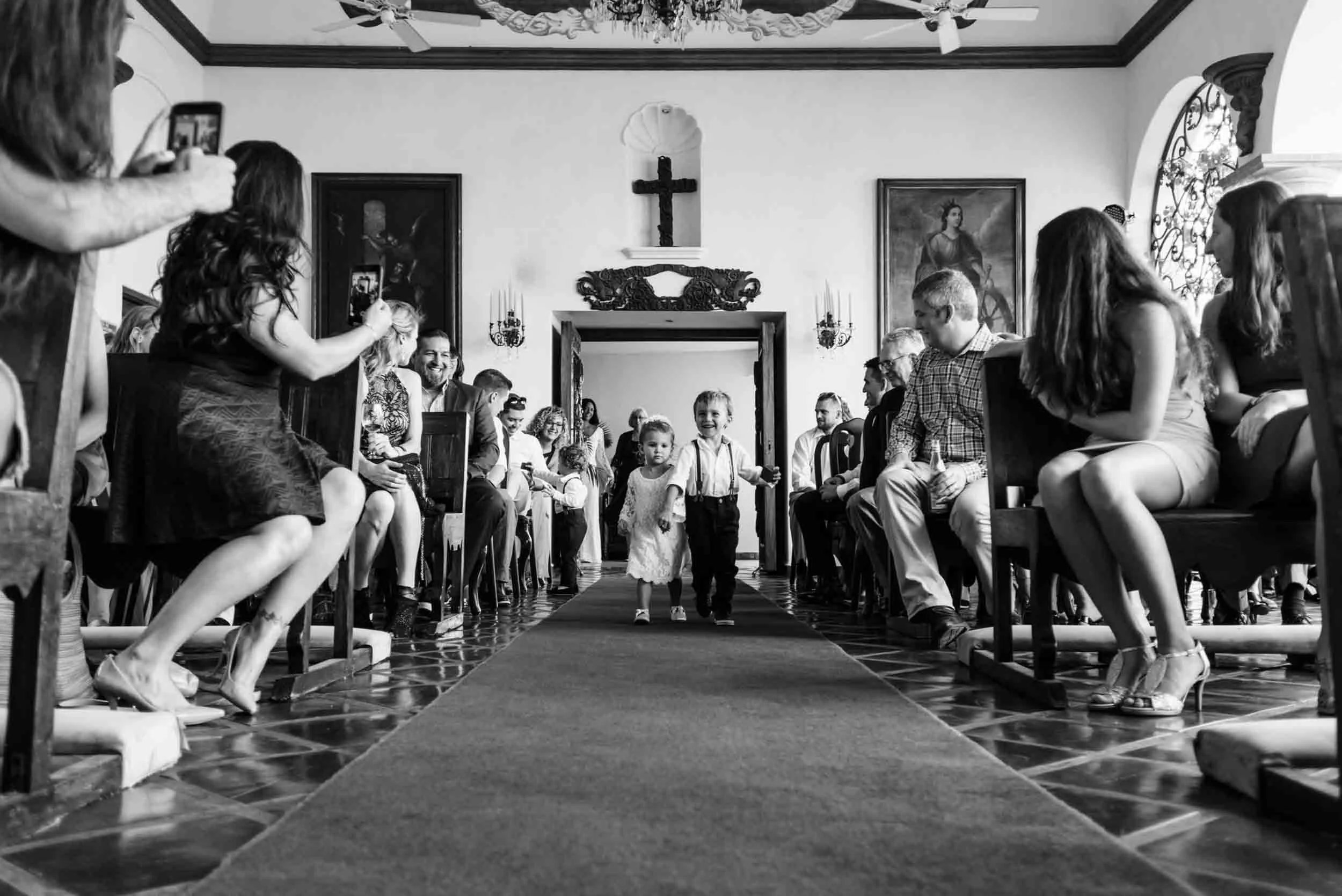Flower girl and ring bearer walk down the aisle of a church during a wedding ceremony, with guests seated on either side, and people taking photos.