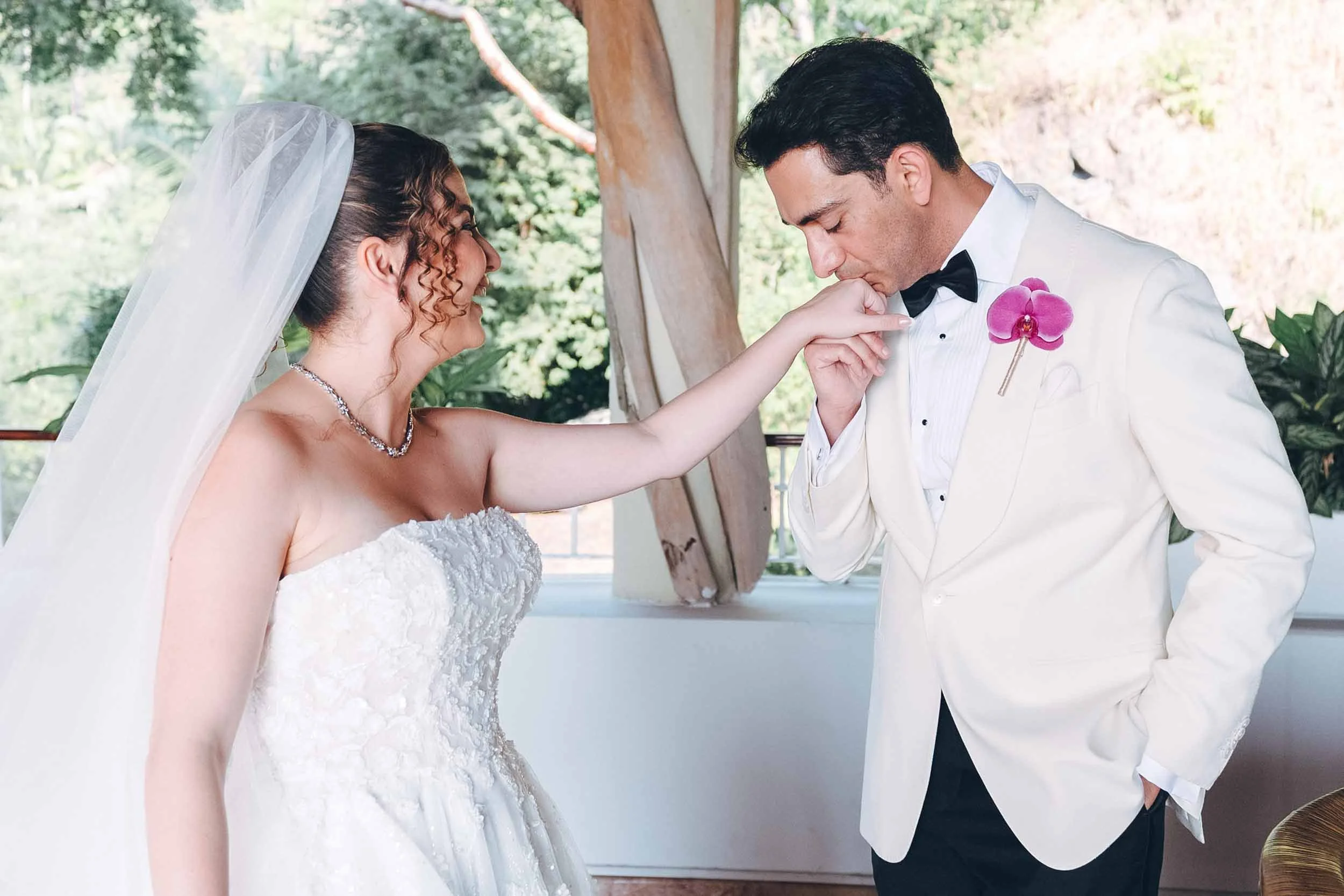 A bride and groom during their first look. The groom is kissing the bride's hand as she smiles.