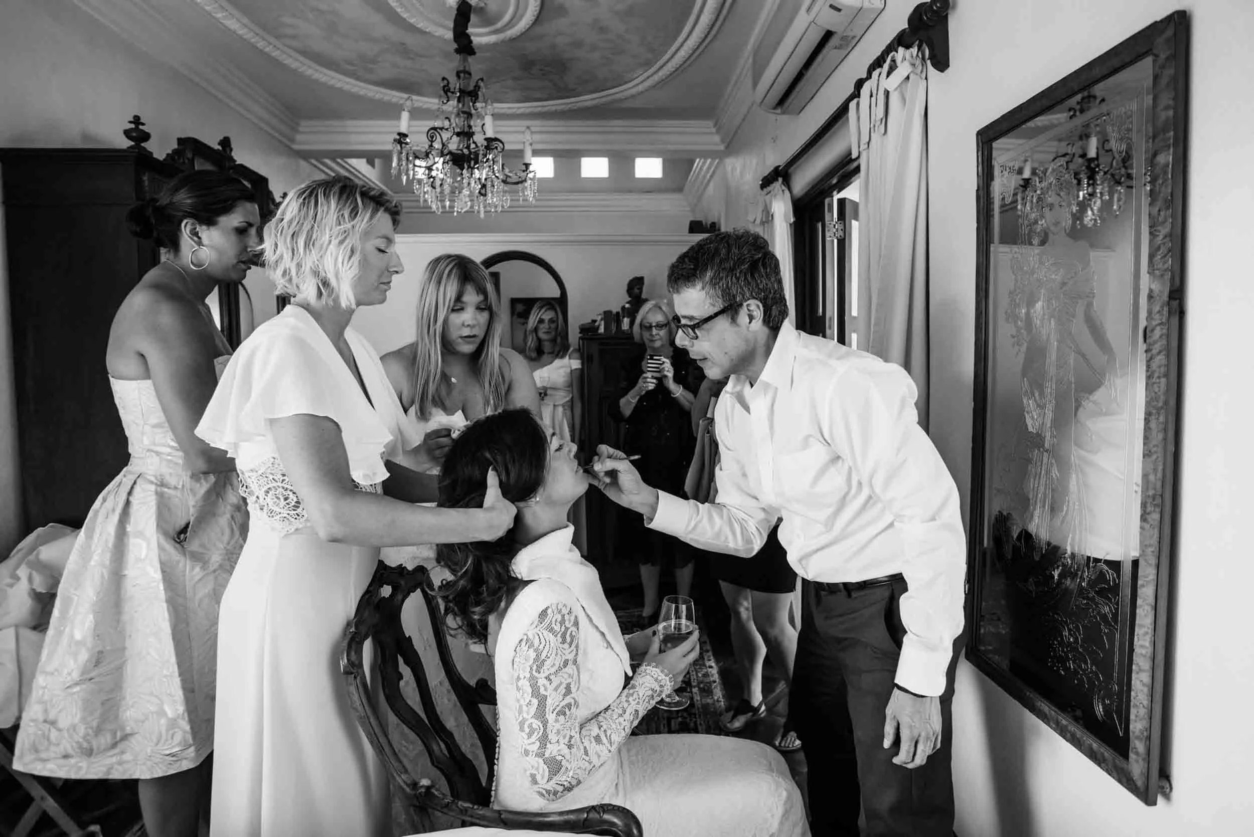 Bride getting makeup done in an elegant room with a chandelier and ornate ceiling. Several women stand around her, some observing and one taking photos. 