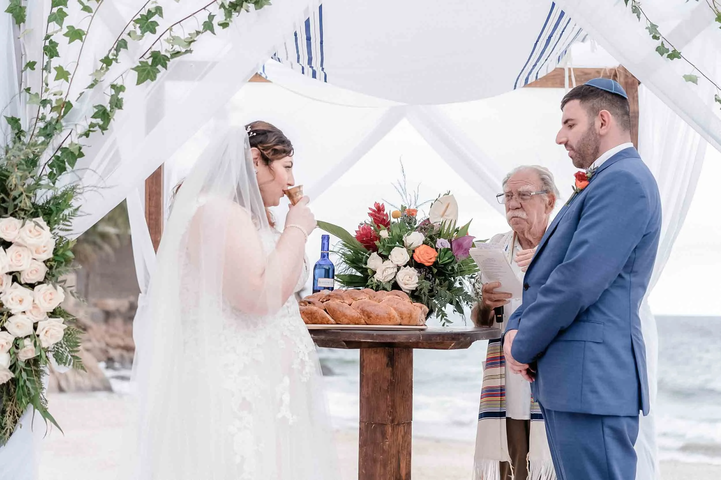 A Jewish wedding ceremony taking place outdoors with a bride and groom standing under a white canopy adorned with flowers, with a rabbi officiating. The bride is eating a piece of bread, and a table with a floral arrangement and challah bread is in f