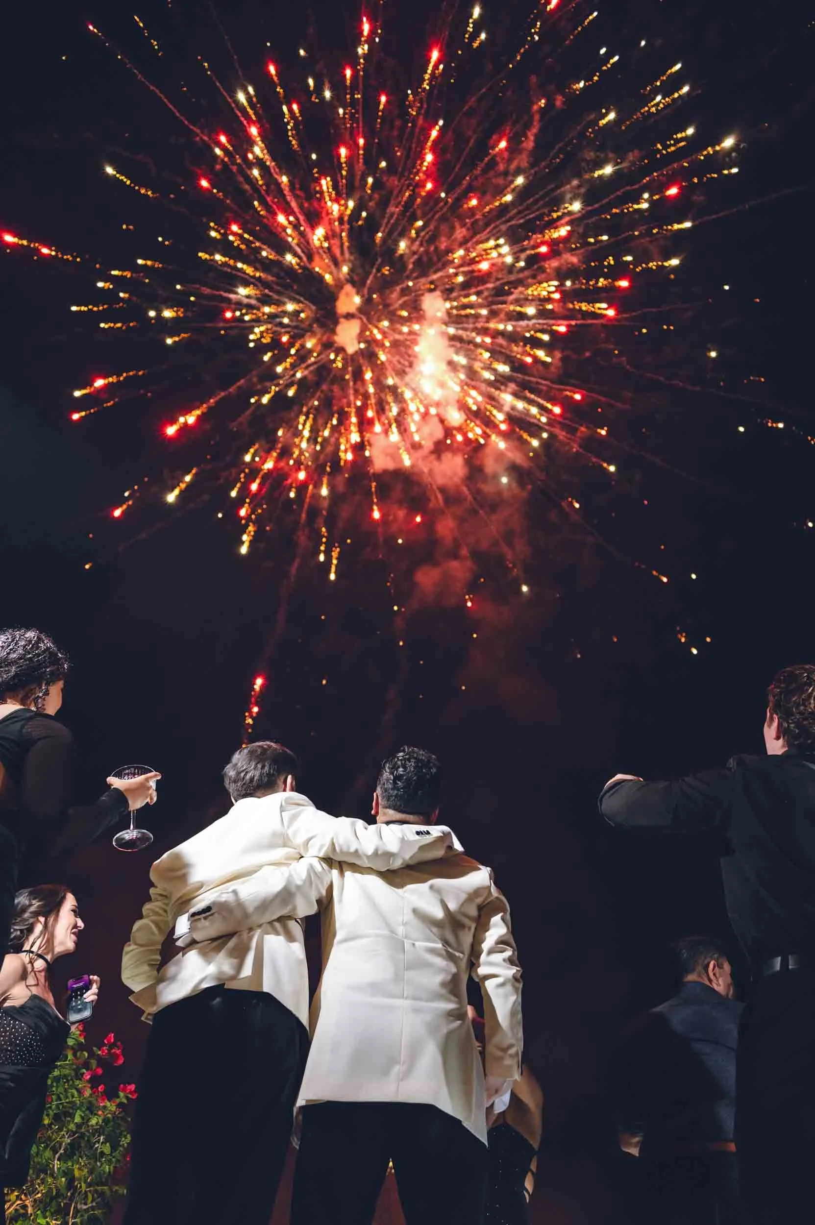 Wedding fireworks in the sky while grooms hug each other on the dance floor along attending guests. 