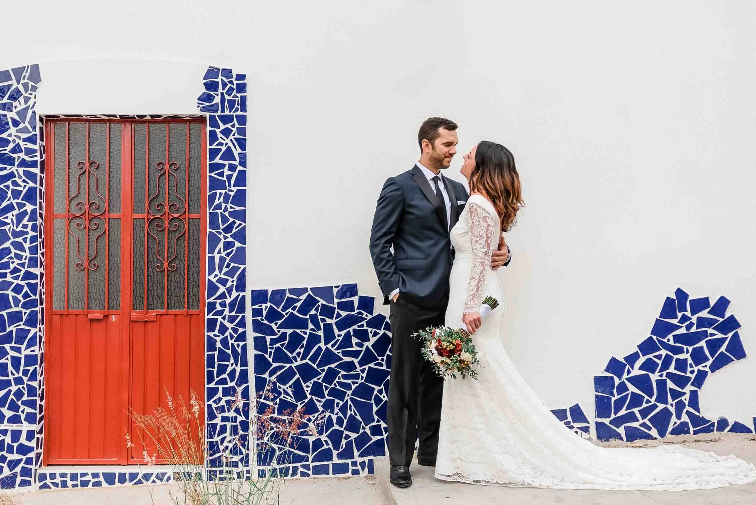 A newlywed couple stands close together outside in front of a white wall with blue mosaic accents and a red door. The groom, dressed in a navy suit, looks at the bride who is wearing a white lace wedding dress and holding a bouquet of flowers. They a
