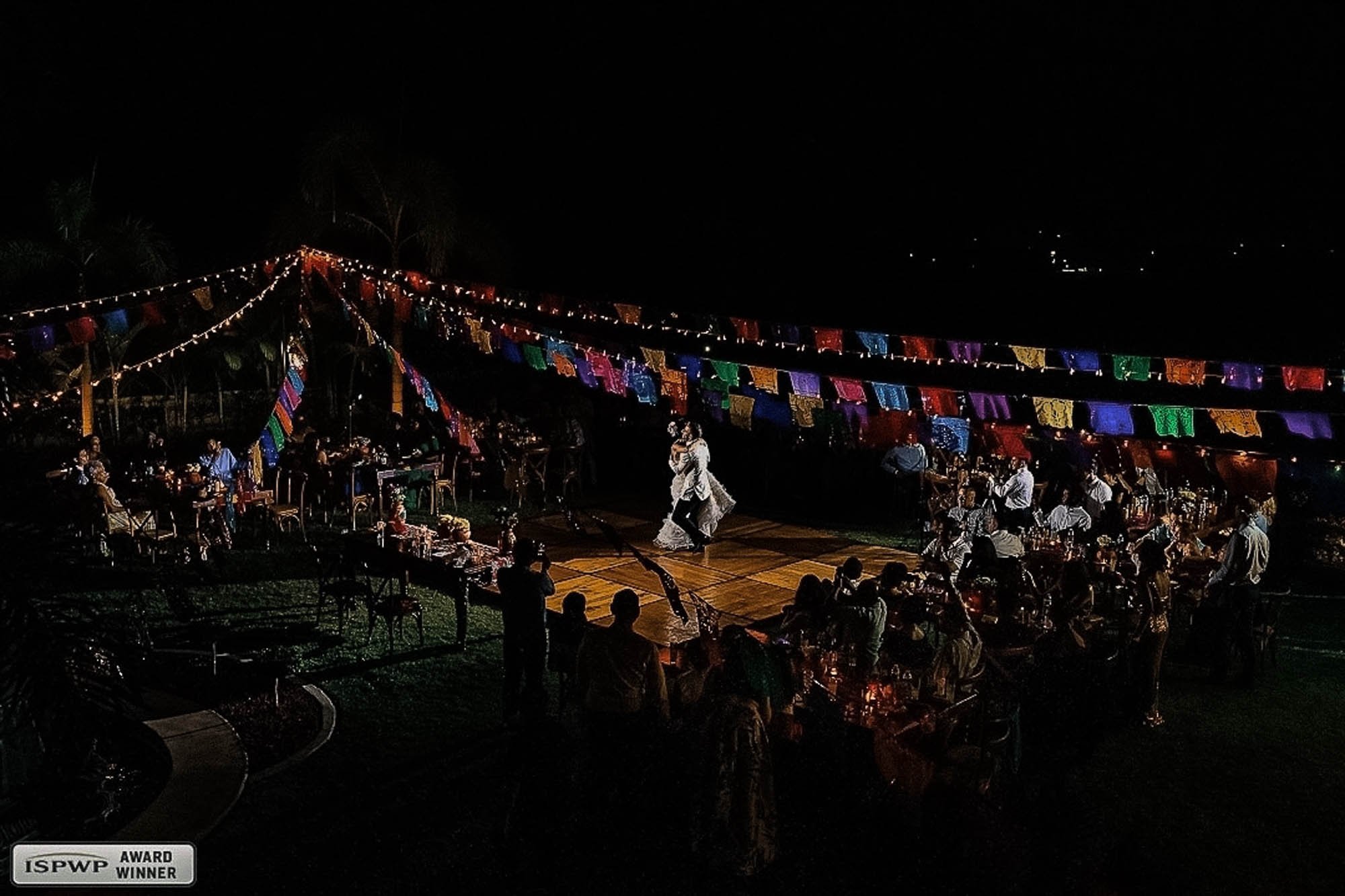 High view of a Mexican wedding with the bride and groom dancing over a papel picado decorated dance floor. 