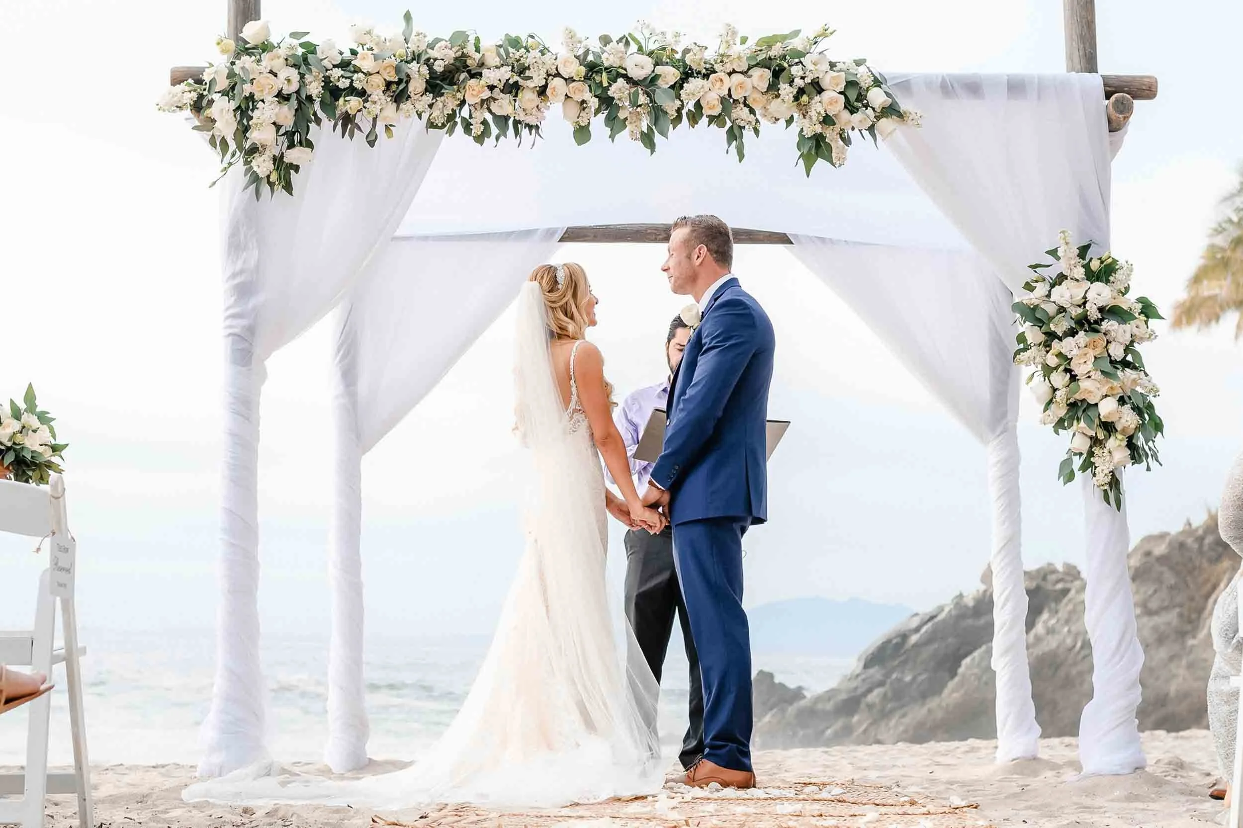 Wedding ceremony on the beach with a bride and groom holding hands and facing each other under a floral and fabric decorated wooden arch, with the ocean and rocky coastline in the background.