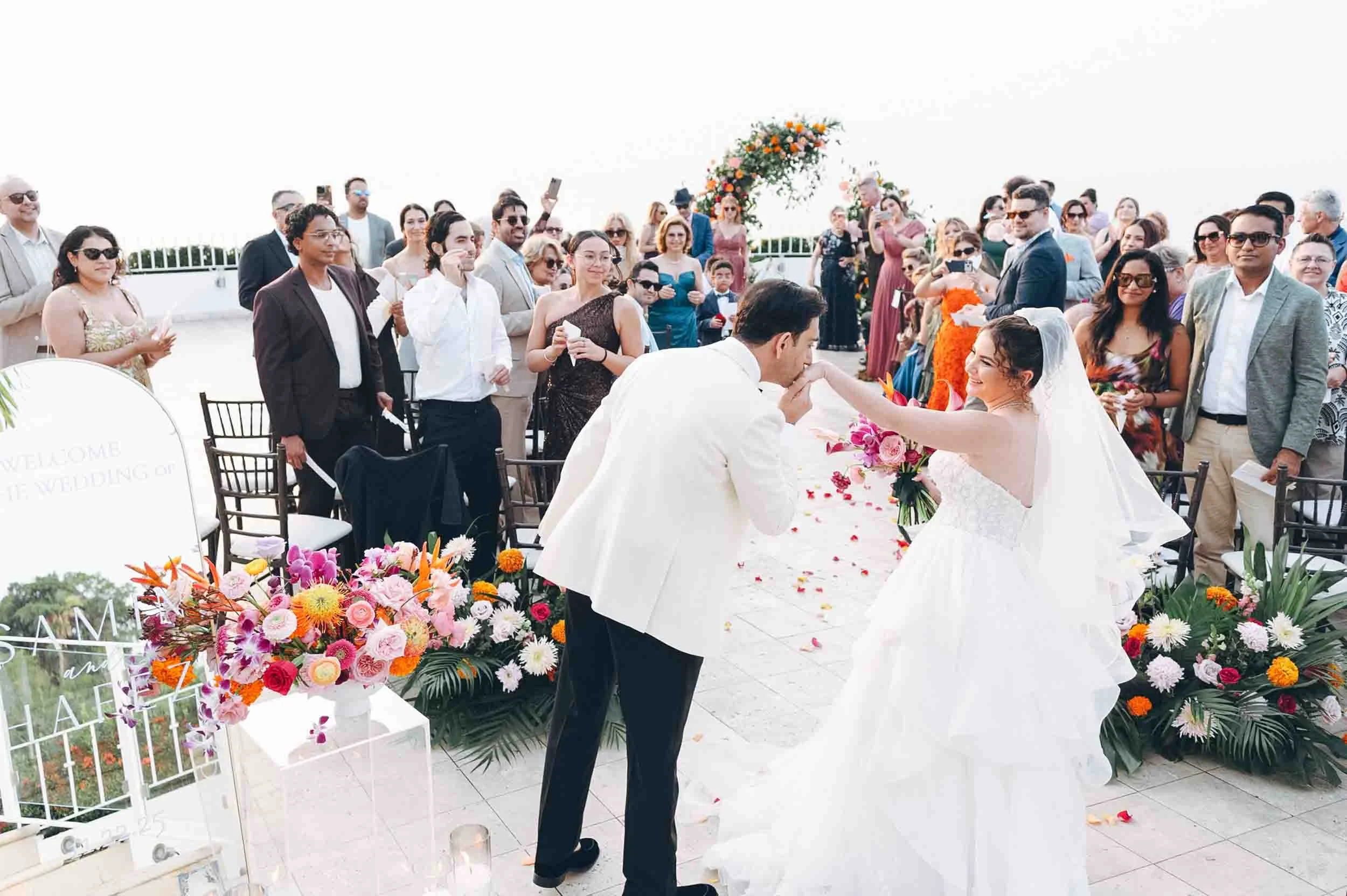 Groom kissing the hand of the bride at their wedding ceremony outdoors, surrounded by seated guests and colorful flowers, with guests taking photos and watching.