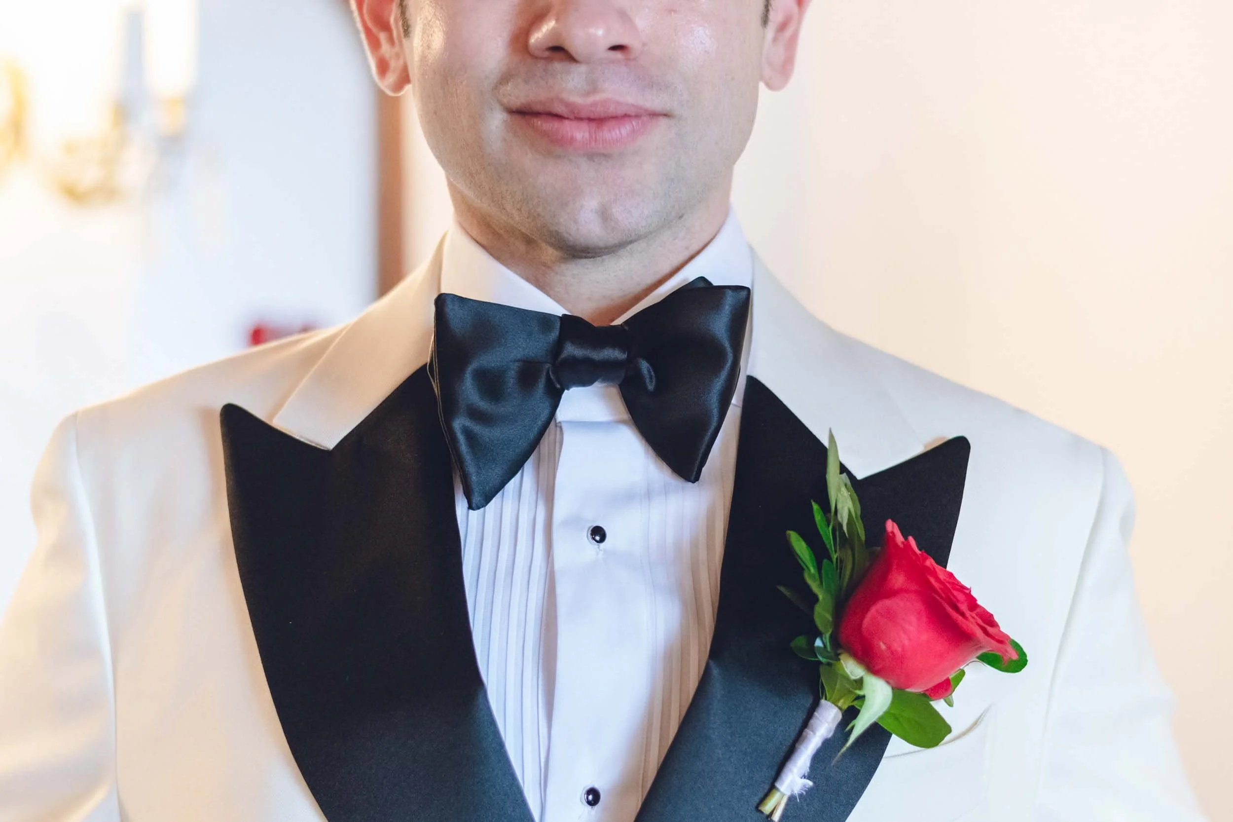 Close-up of a groom wearing a tuxedo with a black bow tie, a white shirt with pinstripes, and a red boutonniere, standing indoors.