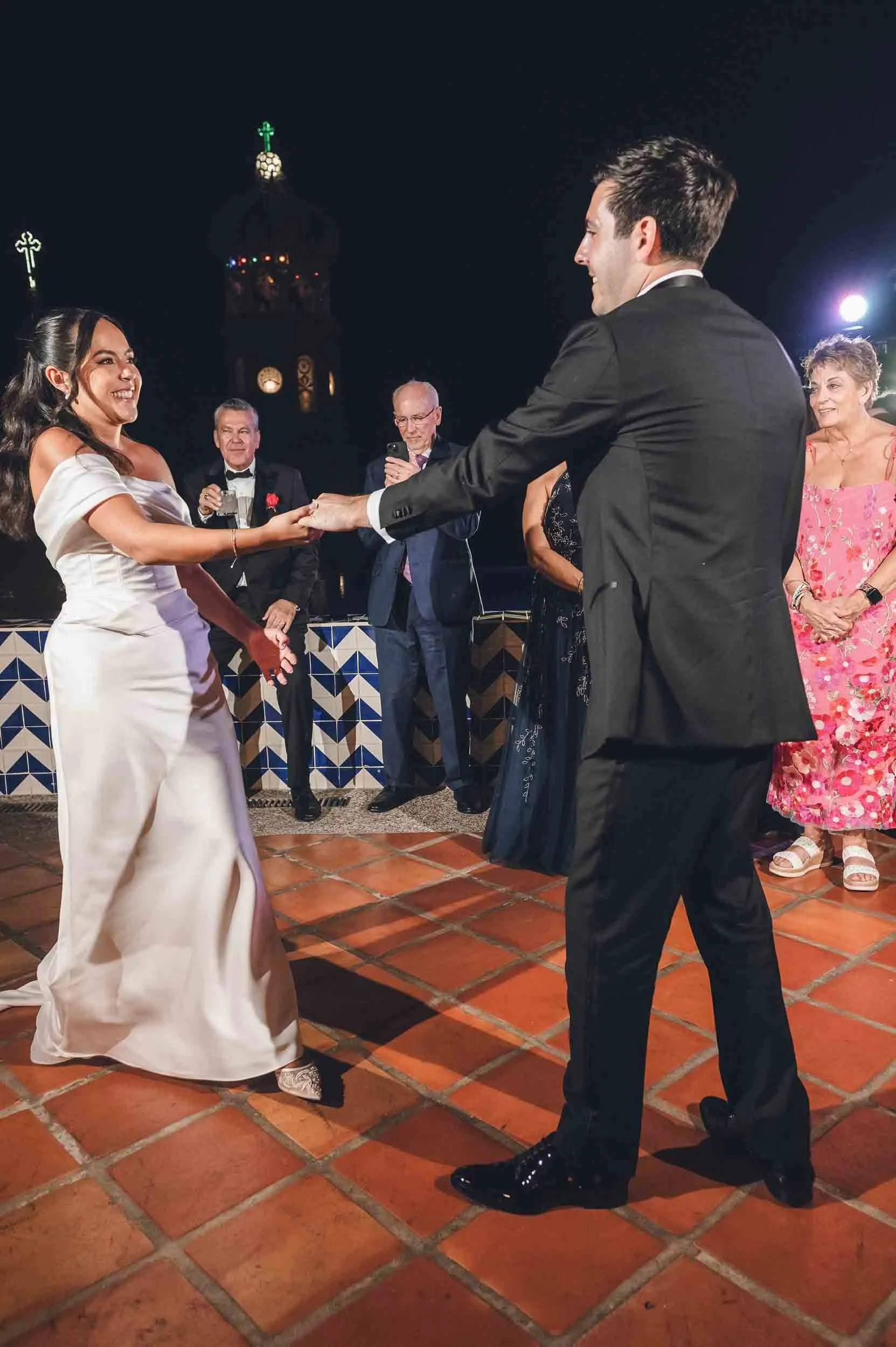 Bride is smiling and holding hands with groom during first dance at night with a historic church in the background.