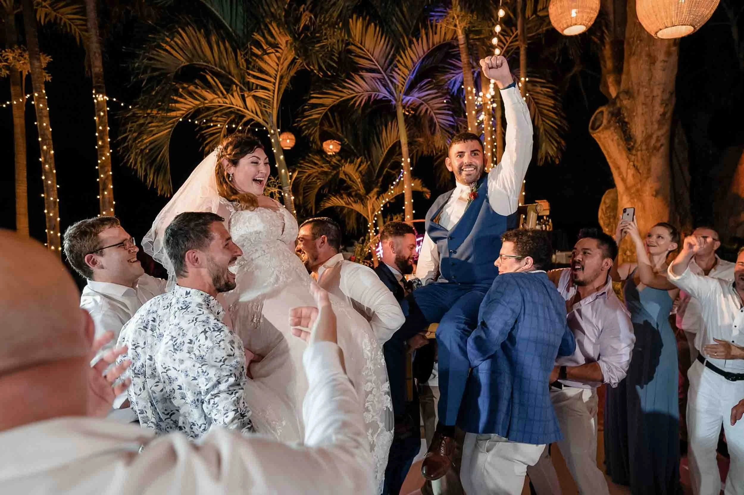 A group of people celebrating at a wedding reception outdoors at night dancing the Hora, with string lights, palm trees, and paper lanterns in the background.