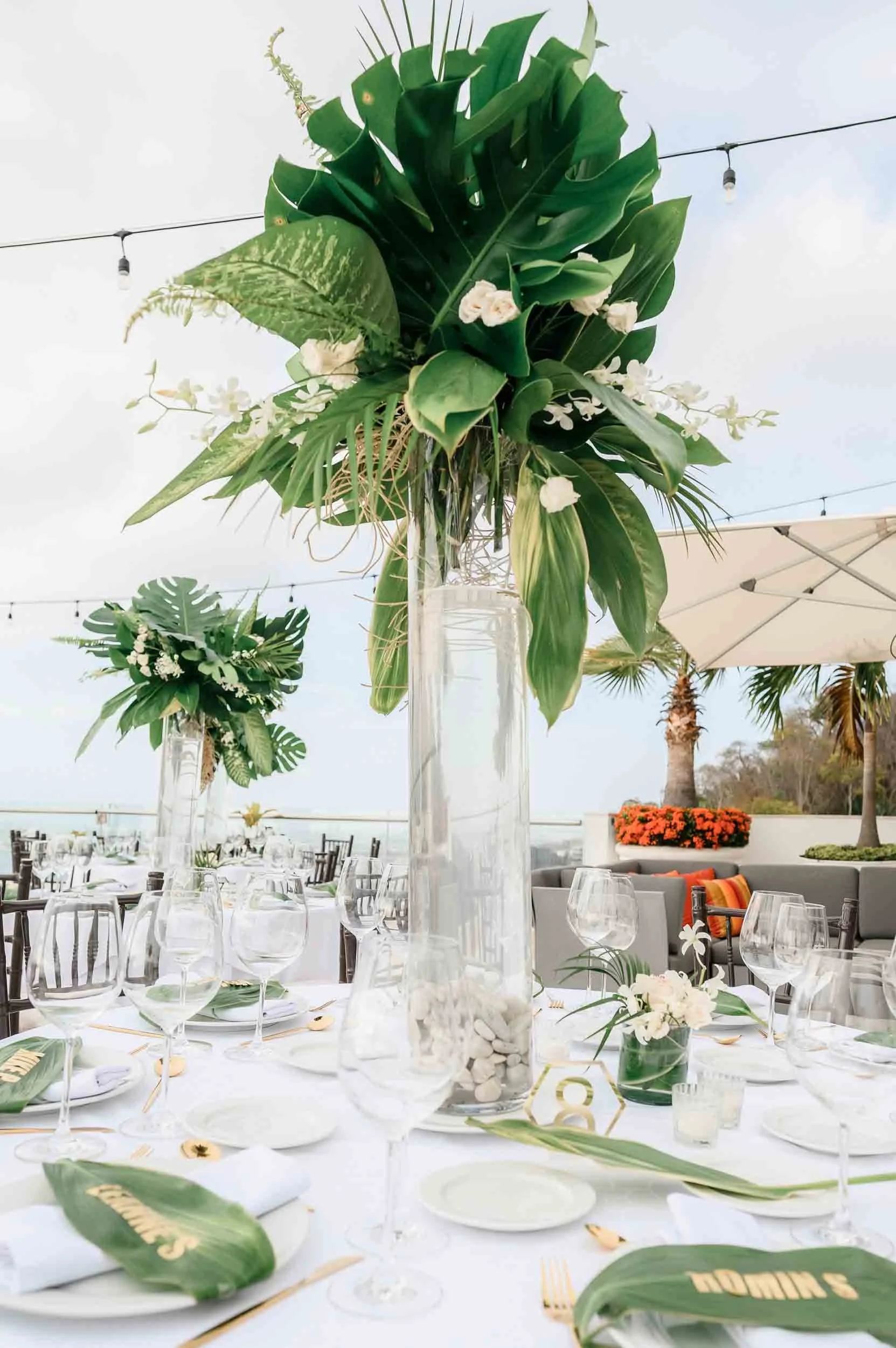 Wedding table setup featuring tall floral arrangements with green leaves and white flowers on a table, with glasses, plates, and decorative elements, set outdoors under a clear sky with string lights and palm trees in the background.
