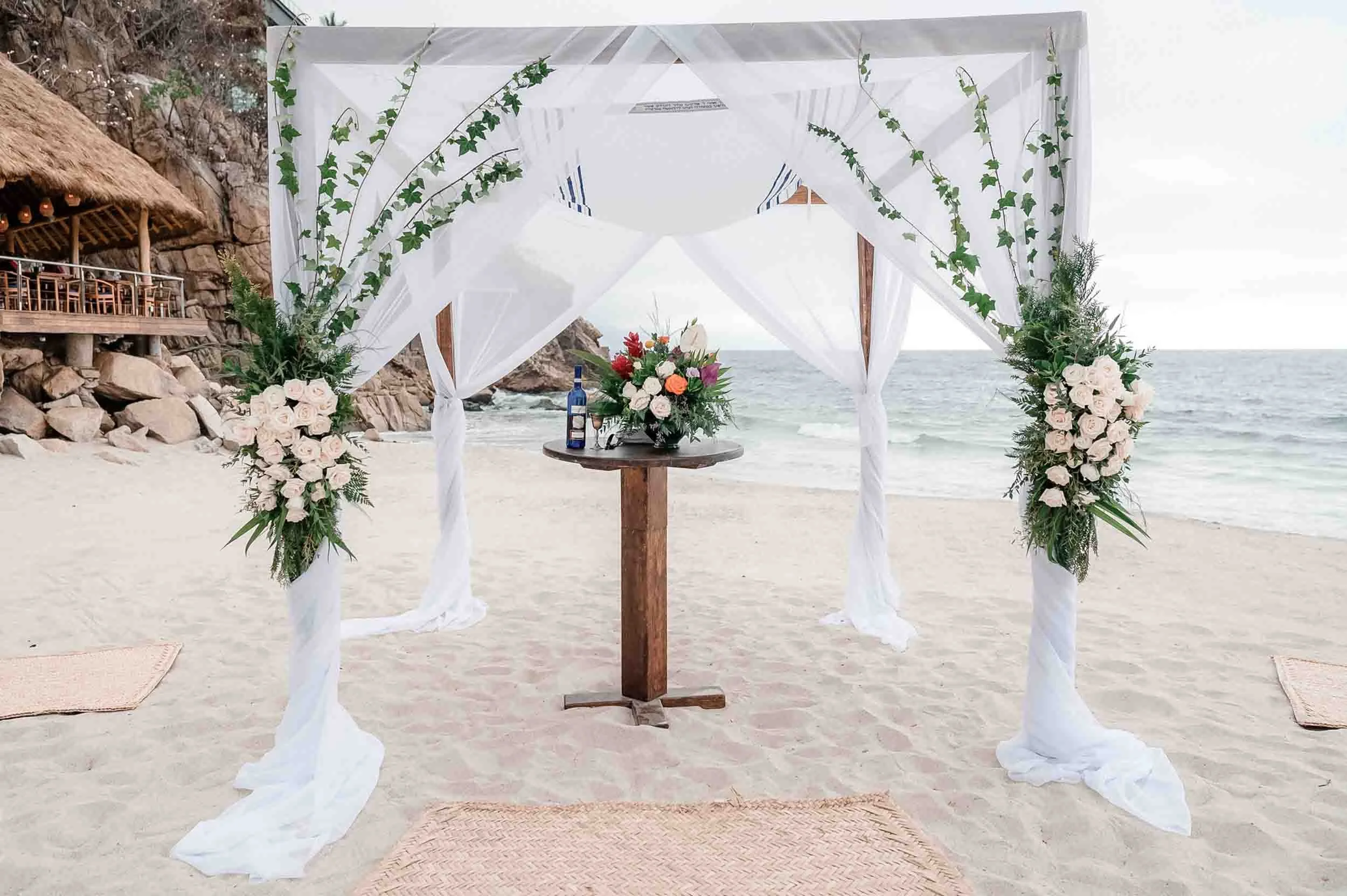 Beach wedding altar with white drapes, floral arrangements, and a table with a bottle of wine, set on the sand facing the ocean.