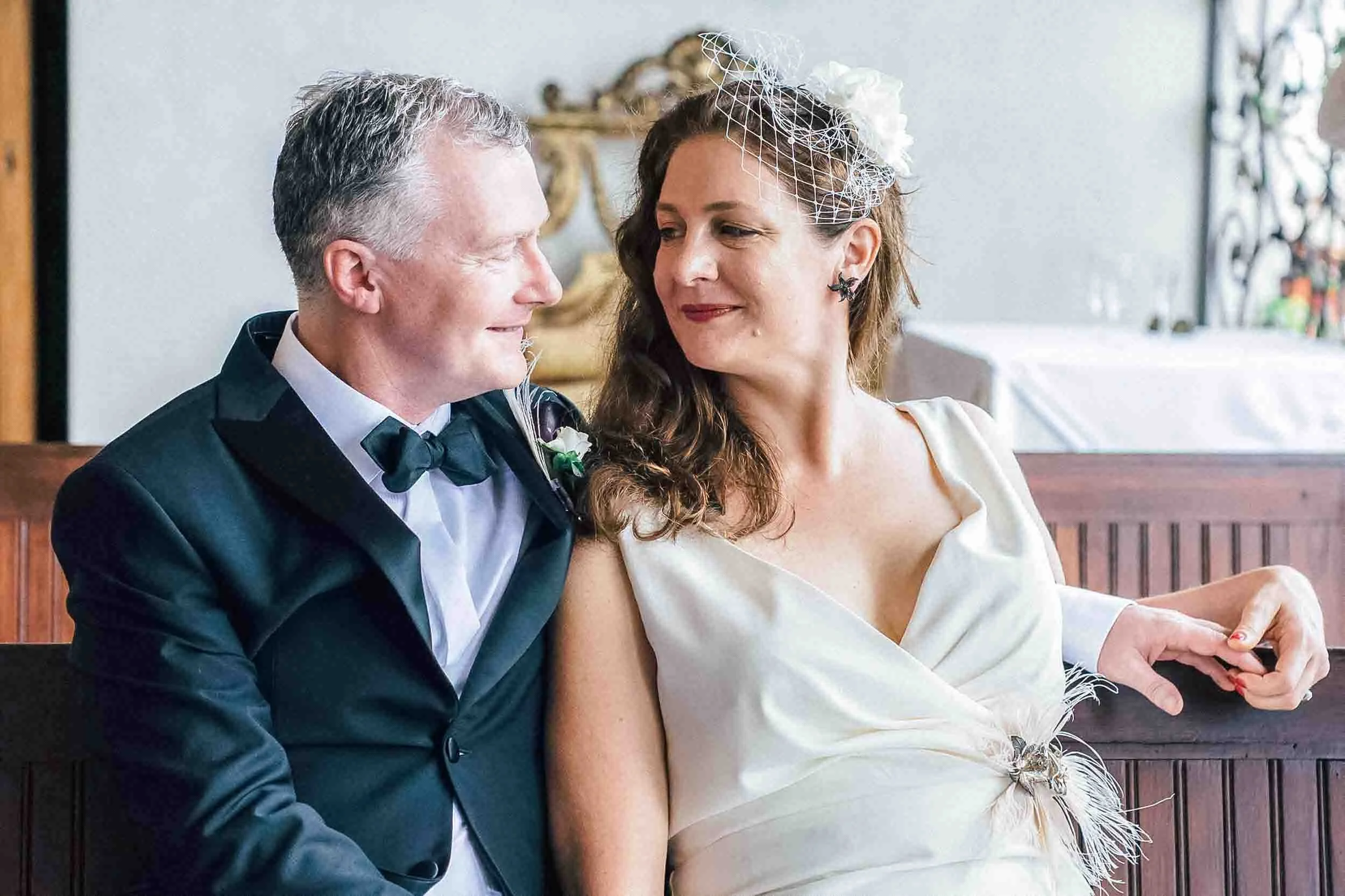 A couple dressed in wedding attire sitting closely together, looking at each other, in a room with a wooden bench and decorative items in the background.