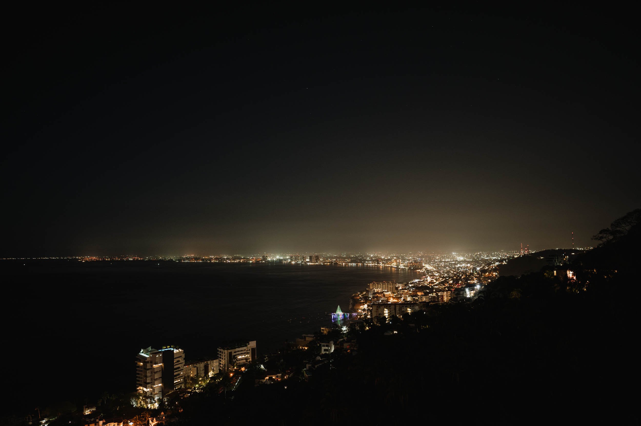 Nighttime cityscape overlooking Puerto Vallarta Mexico with buildings and shoreline lights, under a dark sky.