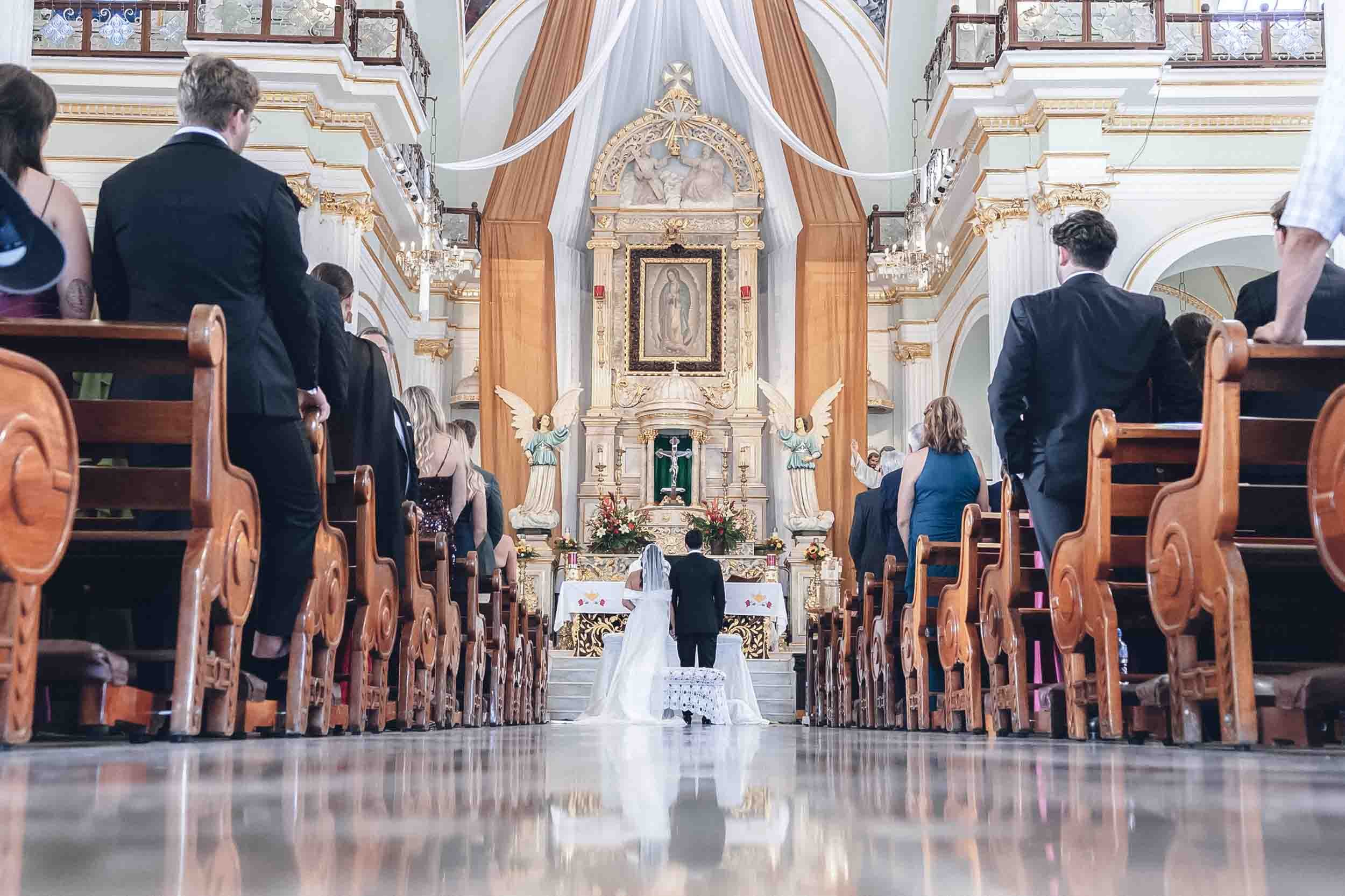 A couple is getting married in a church with guests celebrating. The bride and groom are standing at the altar facing each other, with the bride wearing a white wedding gown and veil. The church has ornate decorations and religious icons at the front