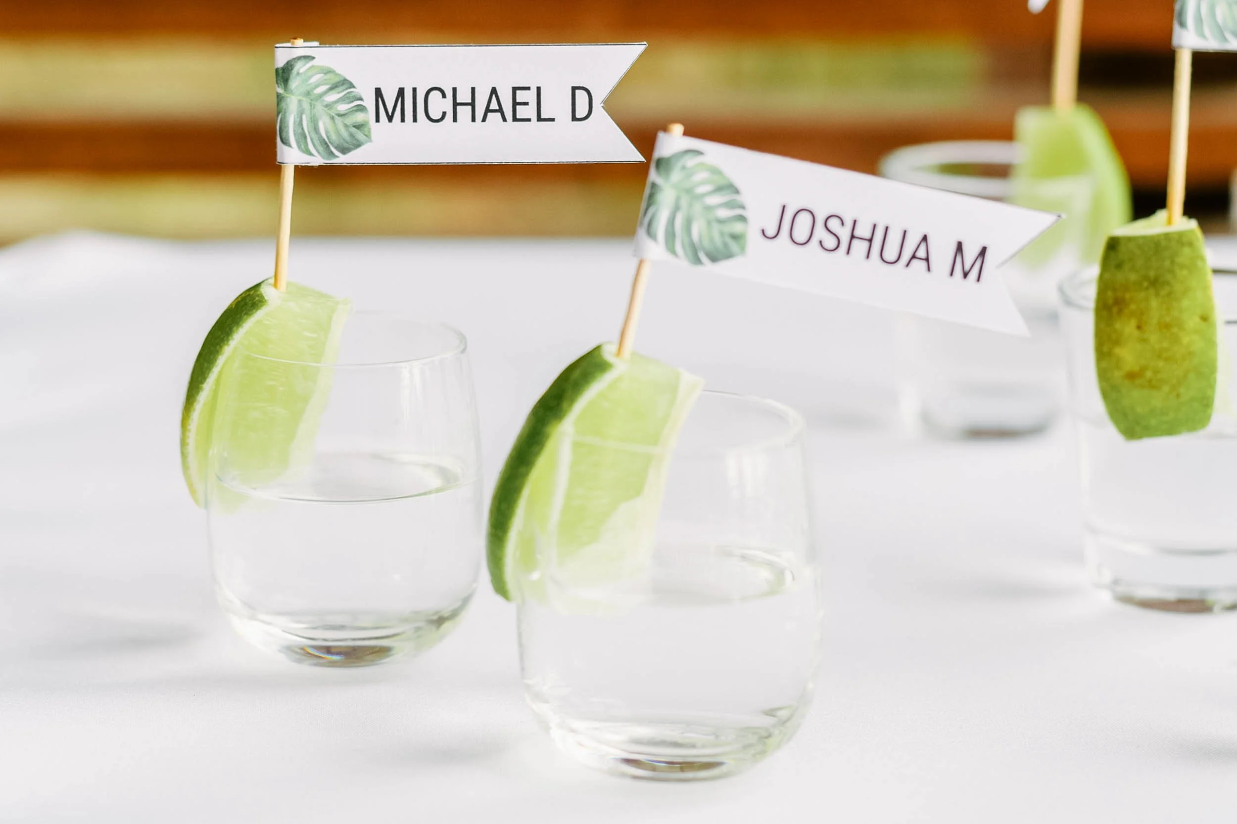 Clear shot of three cocktail glasses with lime wedges and small flags labeled with names, serving as seat assignments at a wedding reception.