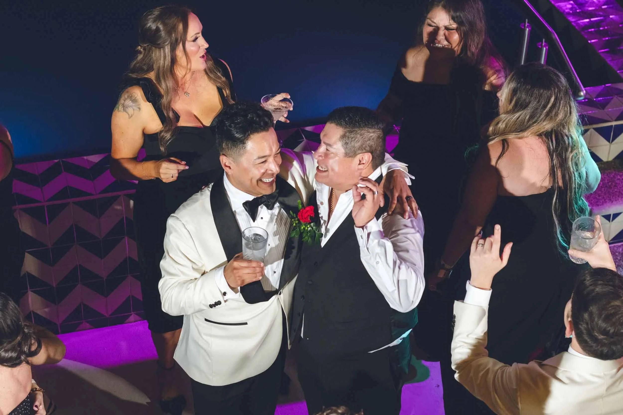 Group of people celebrating at wedding reception  smiling and holding drinks, with a dance floor and vibrant purple lighting in the background.