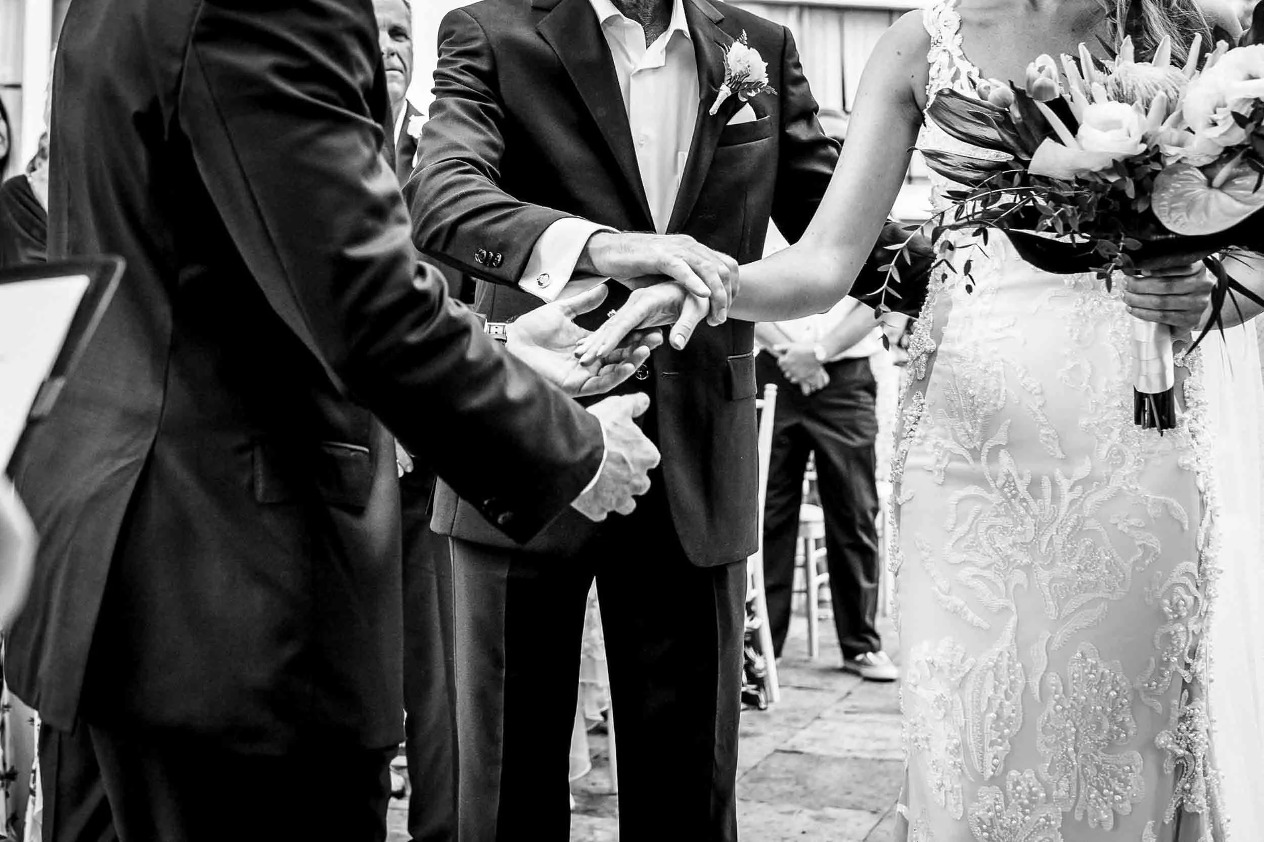 Black and white photo of a wedding ceremony with a bride, groom, and father of the bride giving her hand away. 