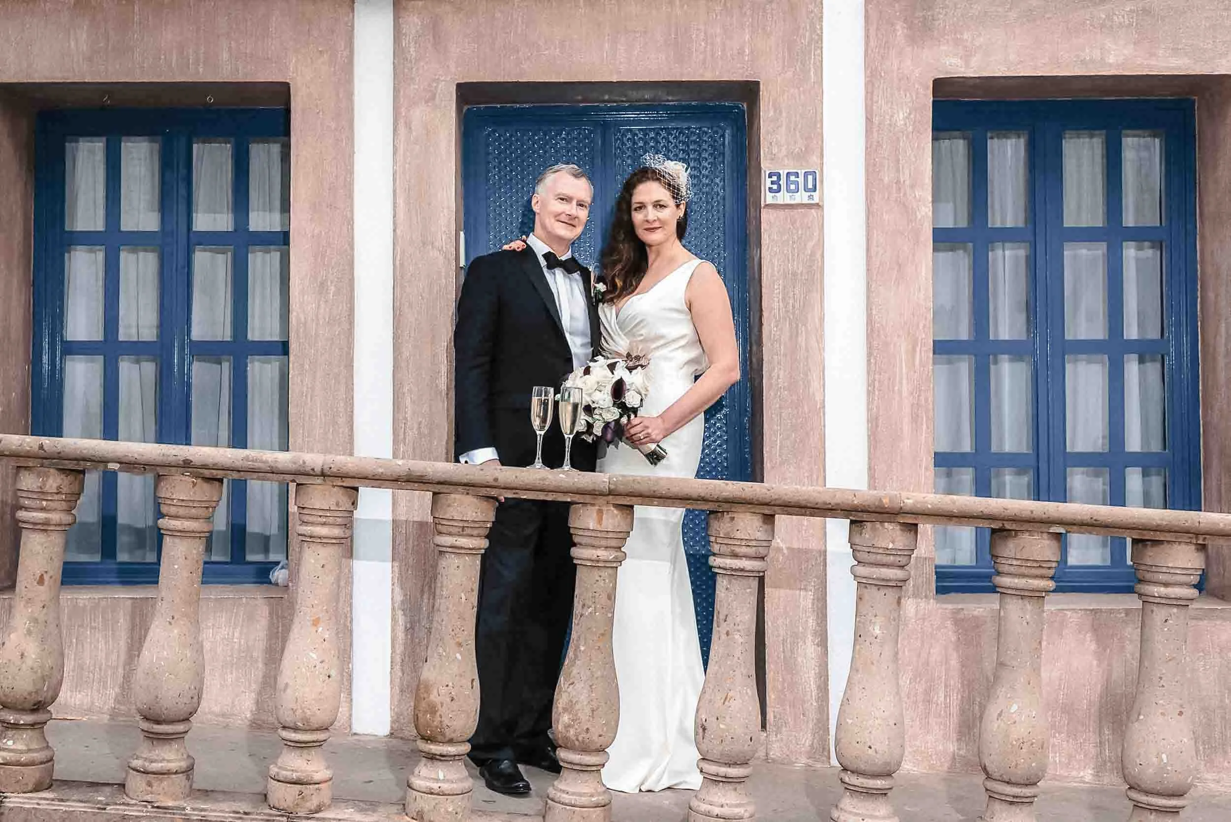 Bride and groom on sidewalk with stone railing. . They smile at the camera in front of a building with blue windows and a blue door in Gringo Gulch.