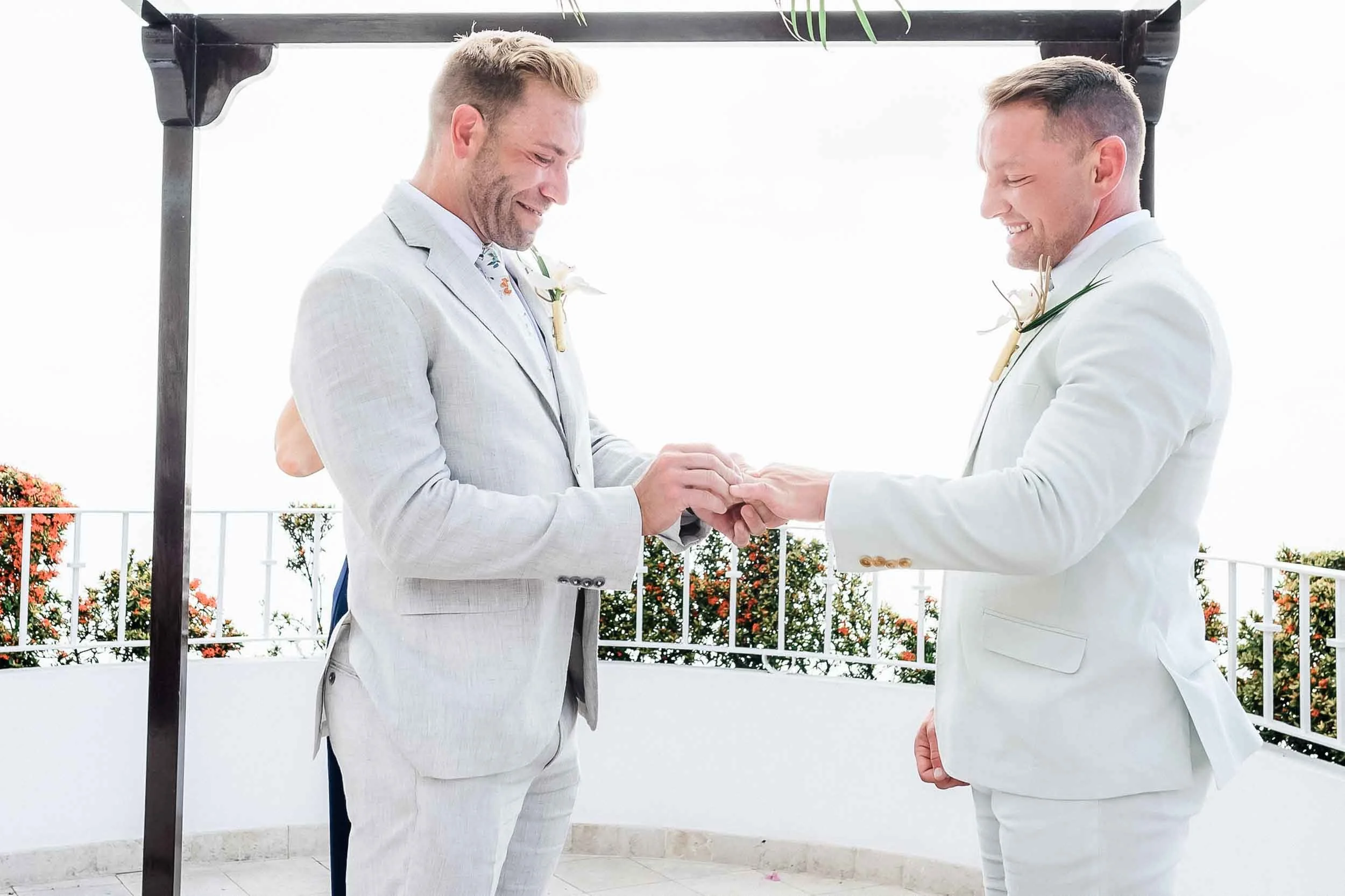 Two men dressed in white suits exchanging rings during a wedding ceremony outdoors under a black arch, with a white railing and potted plants with orange flowers in the background.