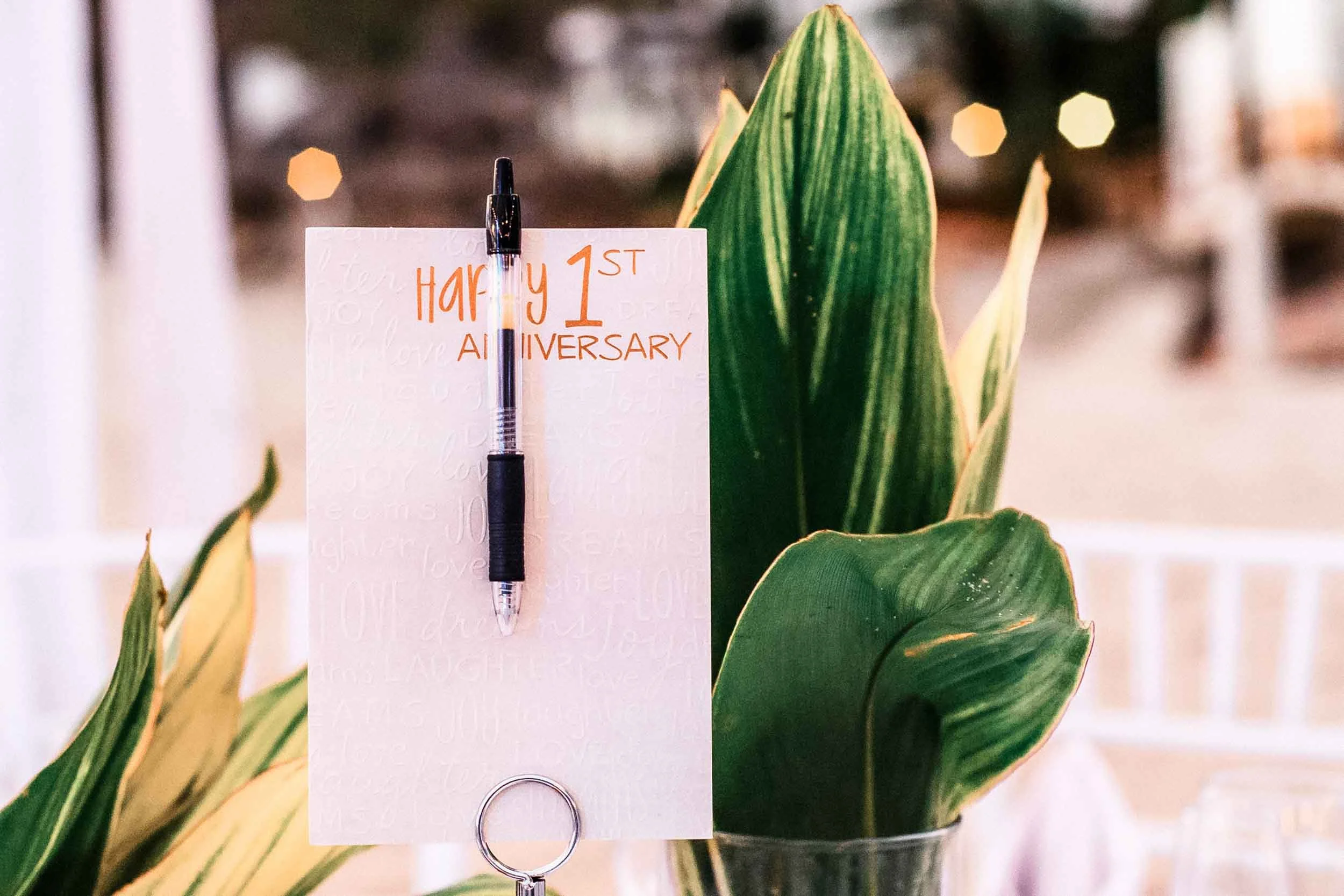 A white card with a handwritten message in orange ink that says "Happy 1st Anniversary" is clipped to a black pen. 