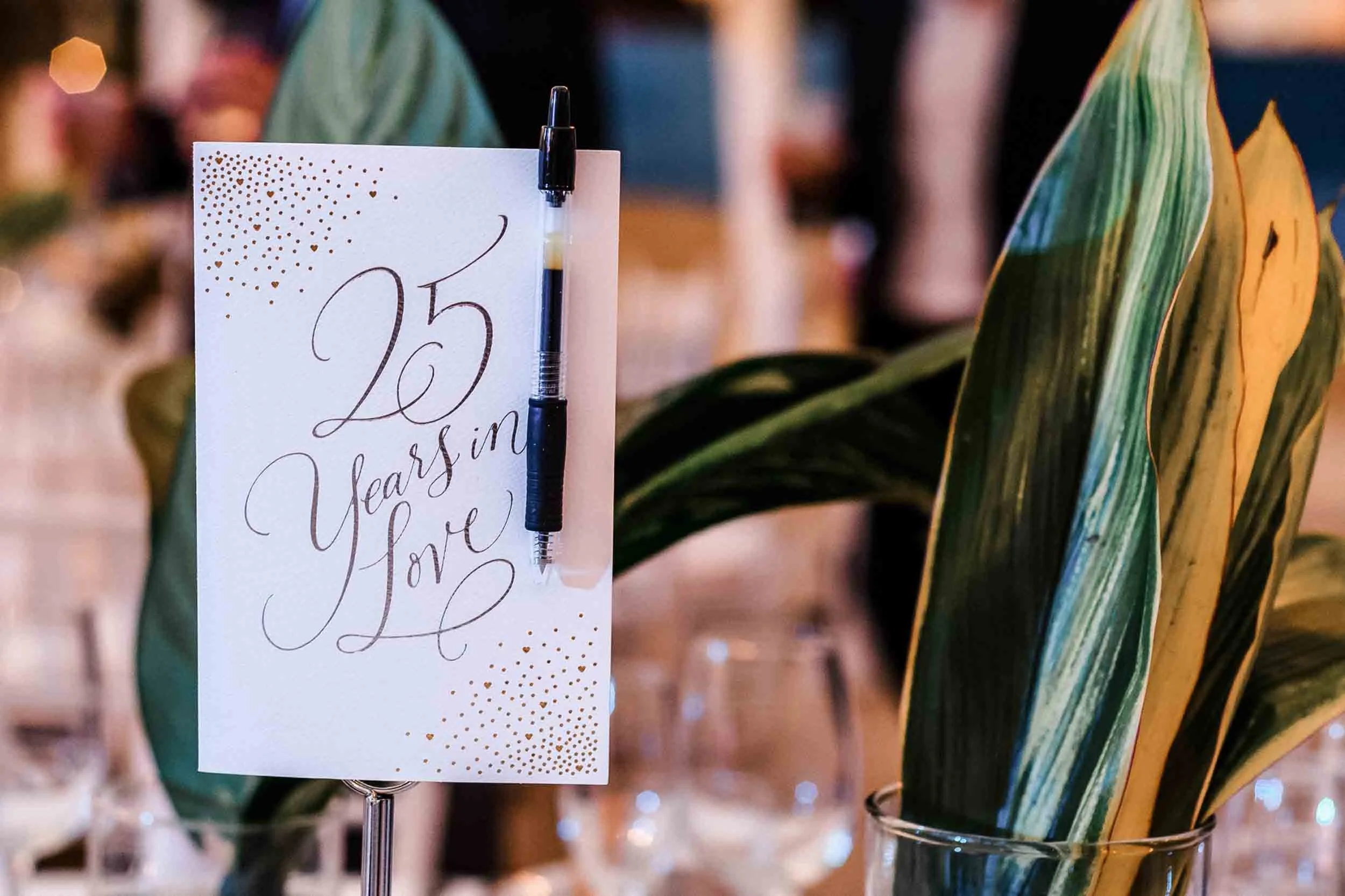 A decorative card attached to a black and clear pen, displayed on a stand, with gold dotted accents and handwritten cursive text, situated next to a large green and yellow variegated plant in a glass container.