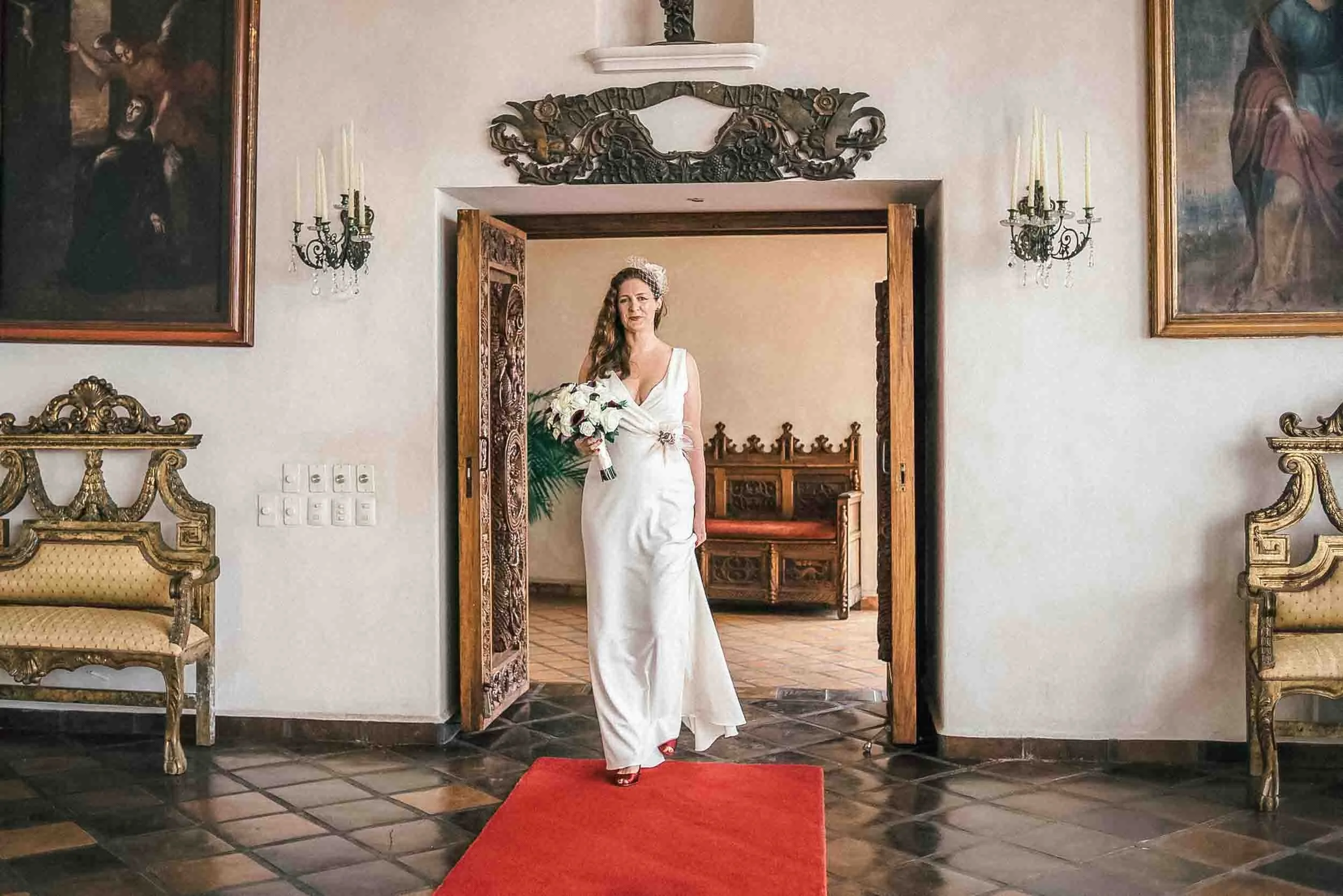  Bride holding a bouquet of white flowers walks on a red carpet towards wedding ceremony.  The room features framed paintings, vintage wall sconces with candles, and decorative chairs with matching gold accents.