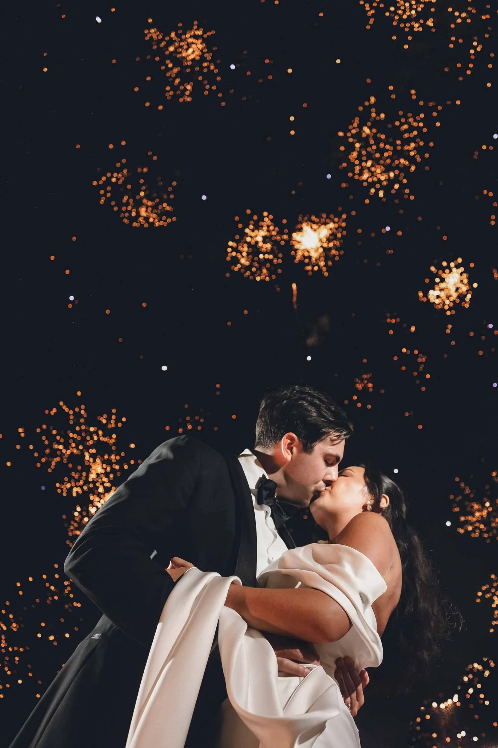 Bride and groom share a romantic kiss during their first dance at their wedding celebration, with fireworks illuminating the night sky behind them.