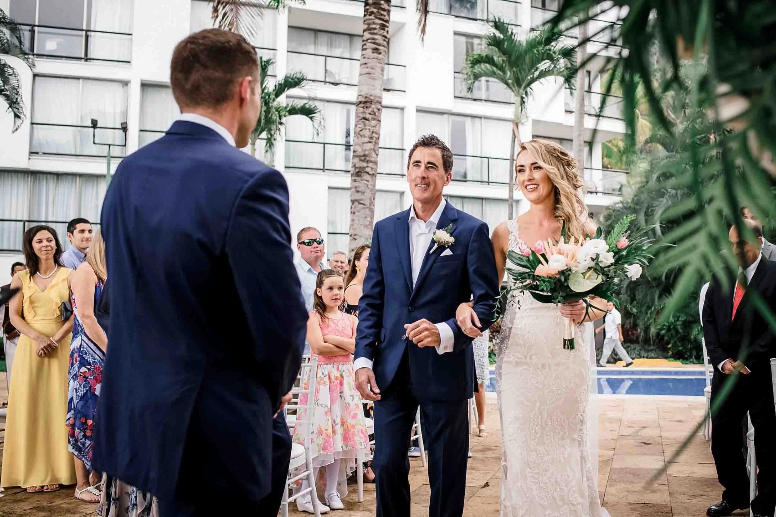 A wedding ceremony outdoors with a bride holding a bouquet of flowers and a groom in a suit, standing facing each other. 