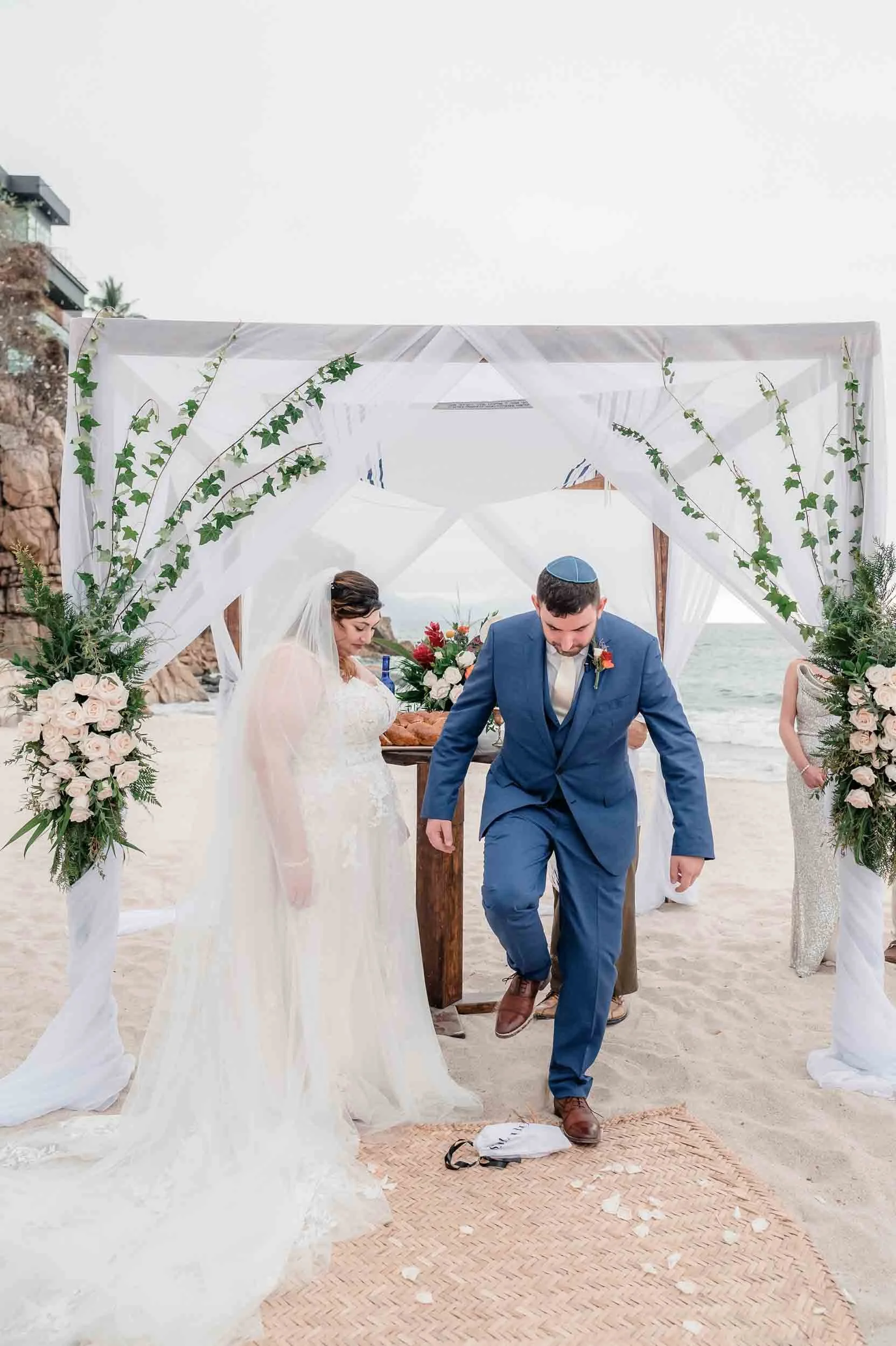 A Jewish beach wedding ceremony with a bride and groom standing under a decorated beach canopy and the groom breaking the glass for Mazel Tov, surrounded by floral arrangements and greenery.