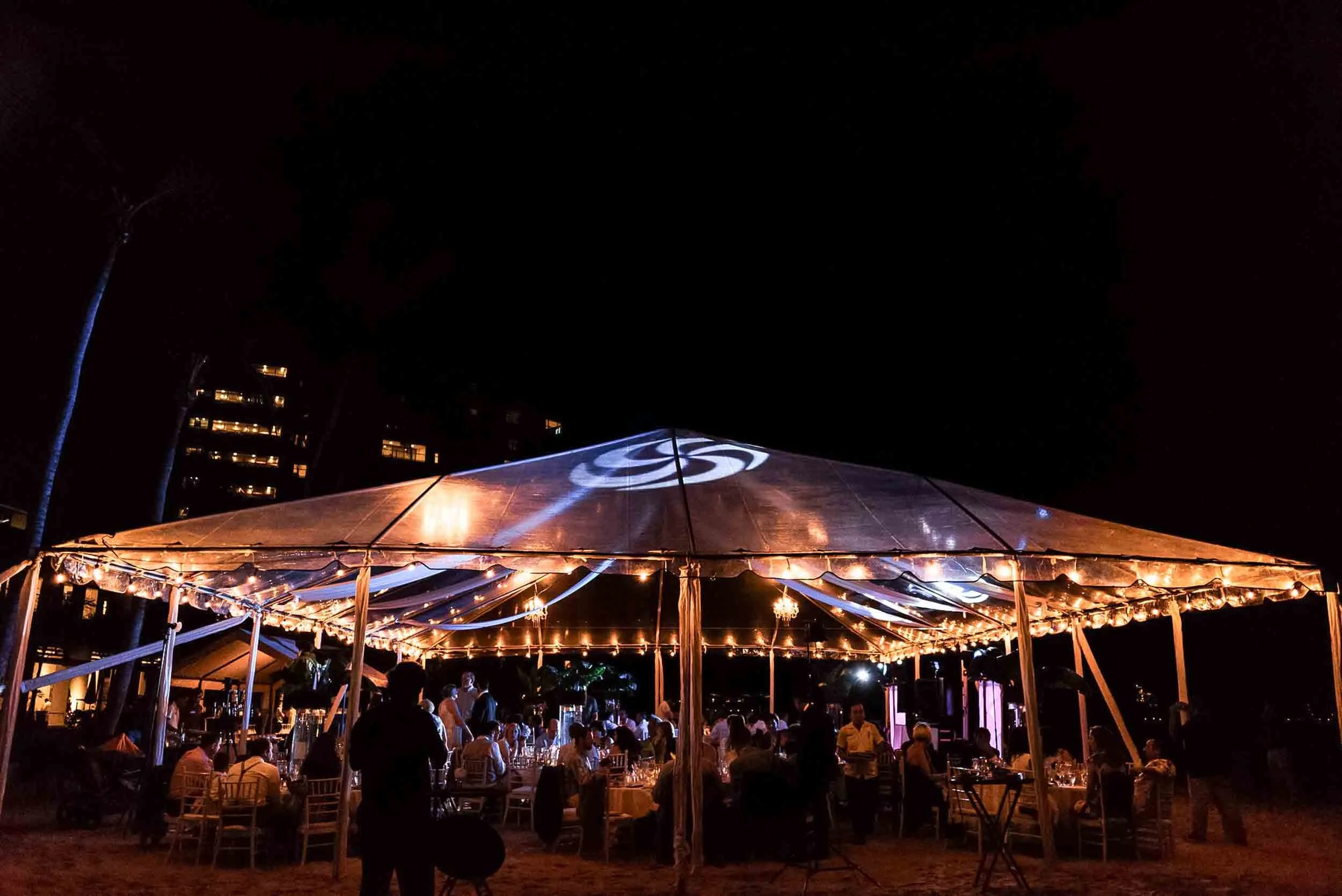 Nighttime outdoor event under a large tent with string lights, surrounded by trees, with people seated and socializing.