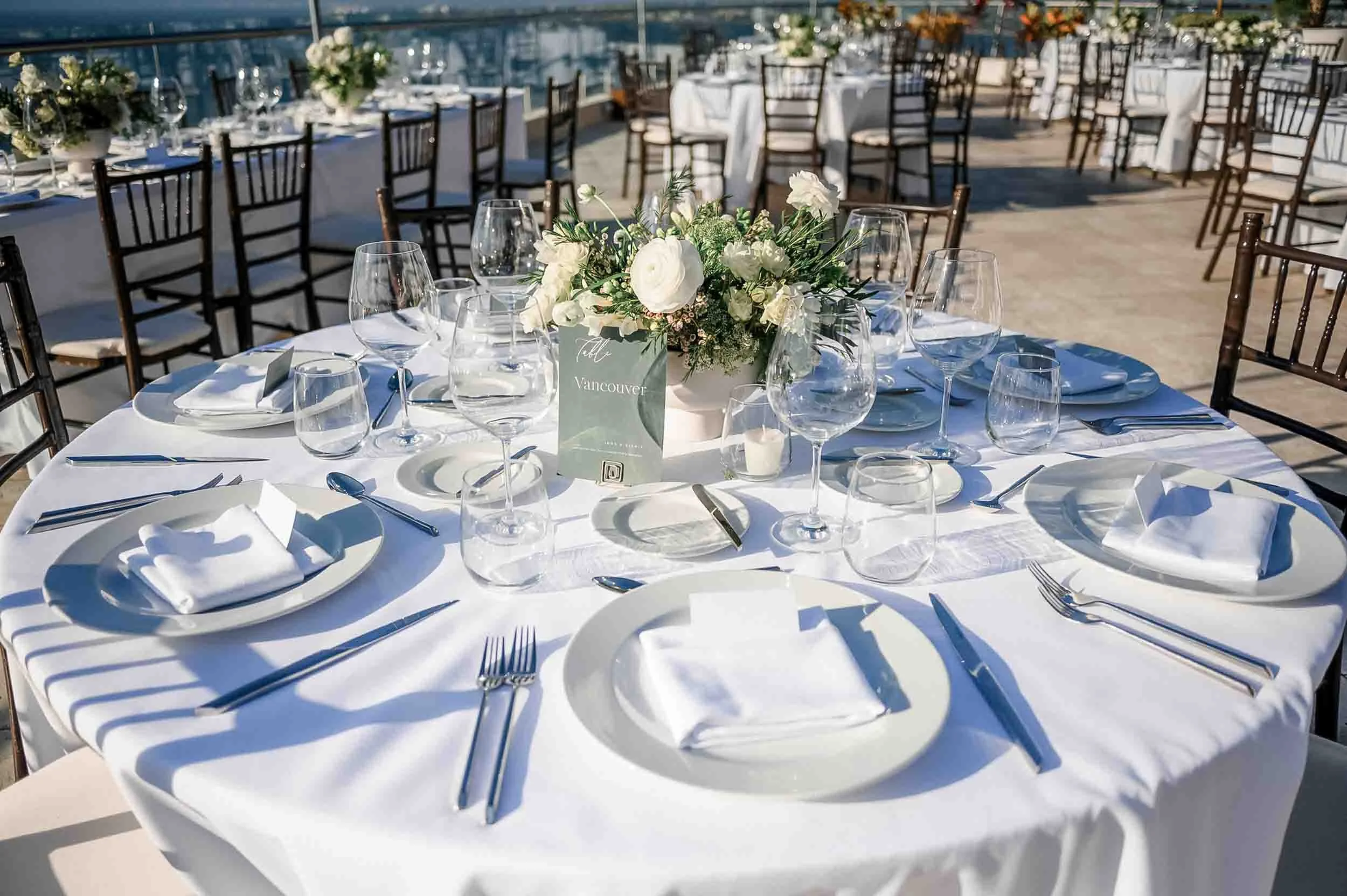 Elegant outdoor wedding reception setup on a rooftop with round tables covered in white tablecloths, decorated with floral centerpieces, glassware, plates, and silverware, overlooking a city skyline.