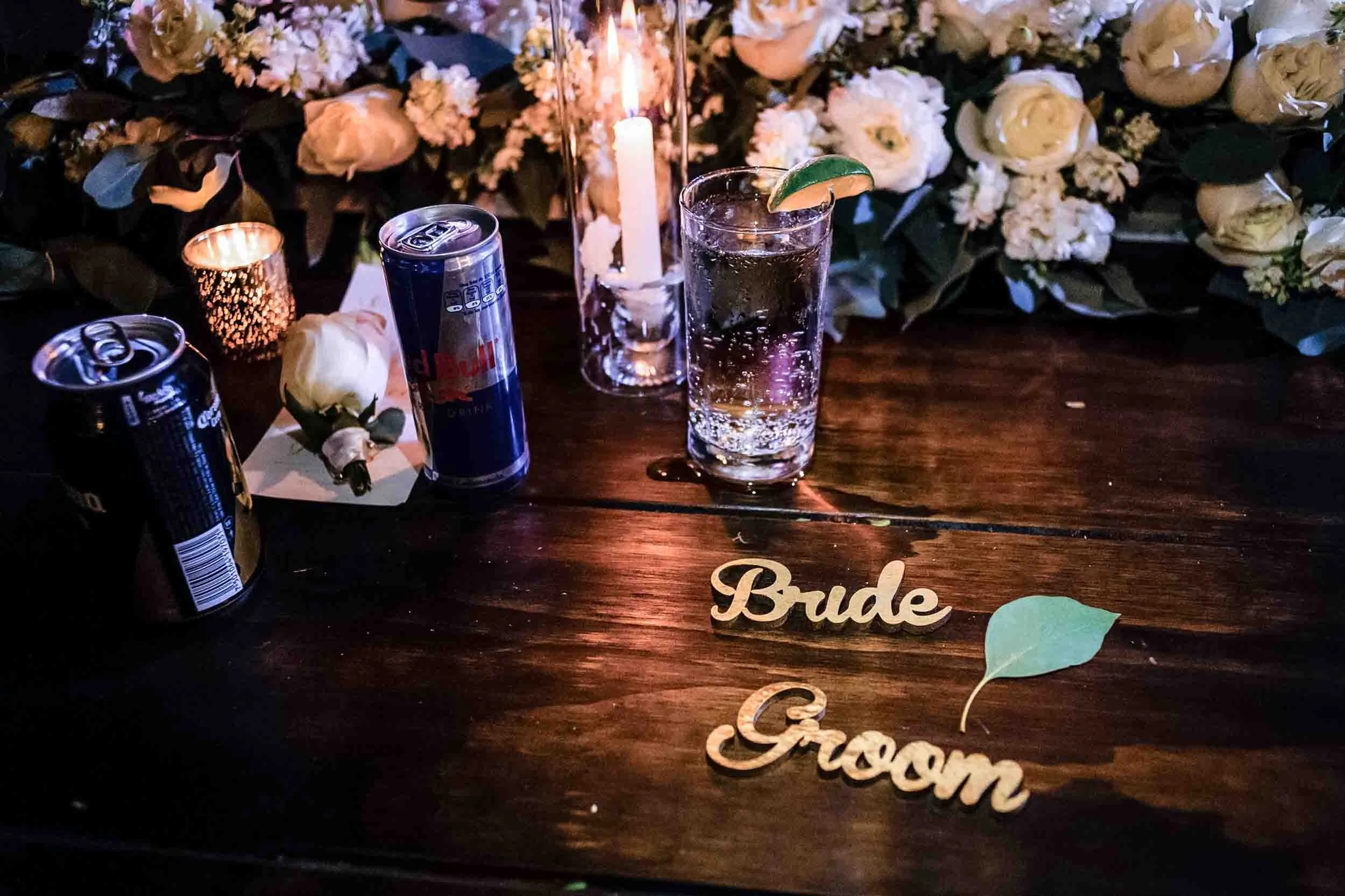 A table decorated with white flowers and greenery, illuminated by candlelight, holding cans of drinks, a glass of water with lime, and a decorative sign that says 'Bride' and 'Groom'.