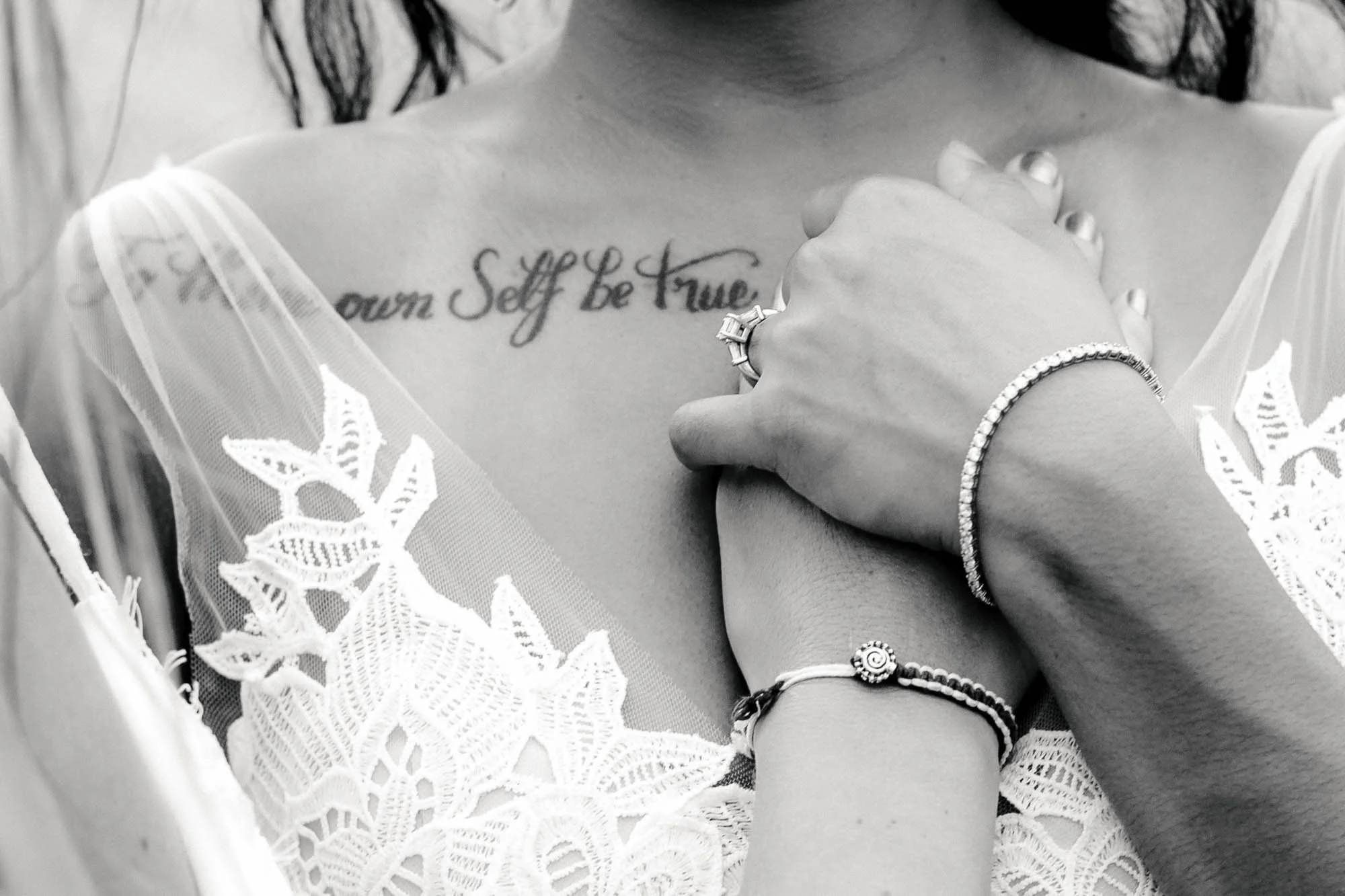 Close-up of  lesbian brides holding hands on the chest of one of them, their hands are next to a tattoo coming from her shoulder.